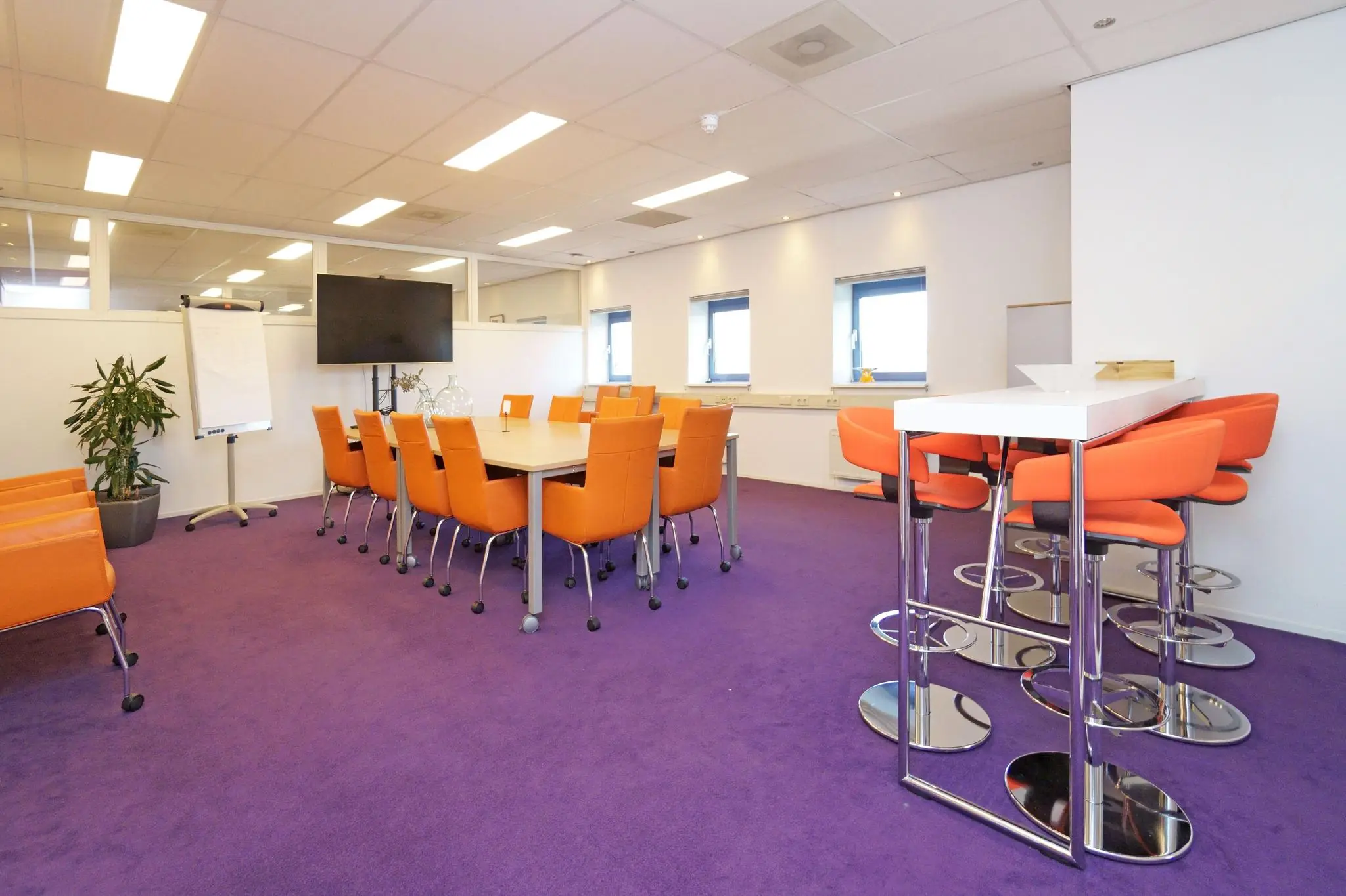 Vergaderruimte aan de Zuiderweg met paarse vloer, oranje stoelen, een grote tafel, een flatscreen en hoge tafel met barkrukken.