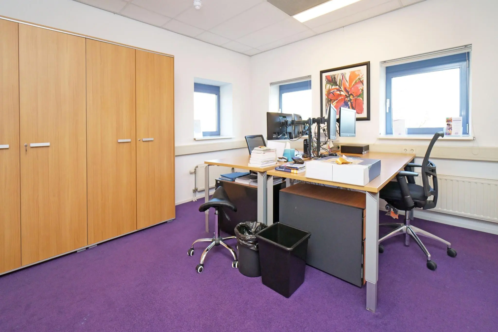 Ingericht kantoor met paarse vloerbedekking, houten kasten en twee werkplekken met bureaus en computerschermen.