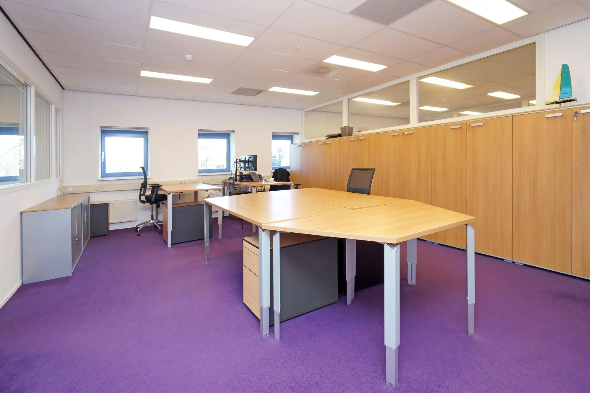 Kantoorruimte aan de Zuiderweg met paarse vloerbedekking, houten bureaus en ingebouwde opbergkasten.