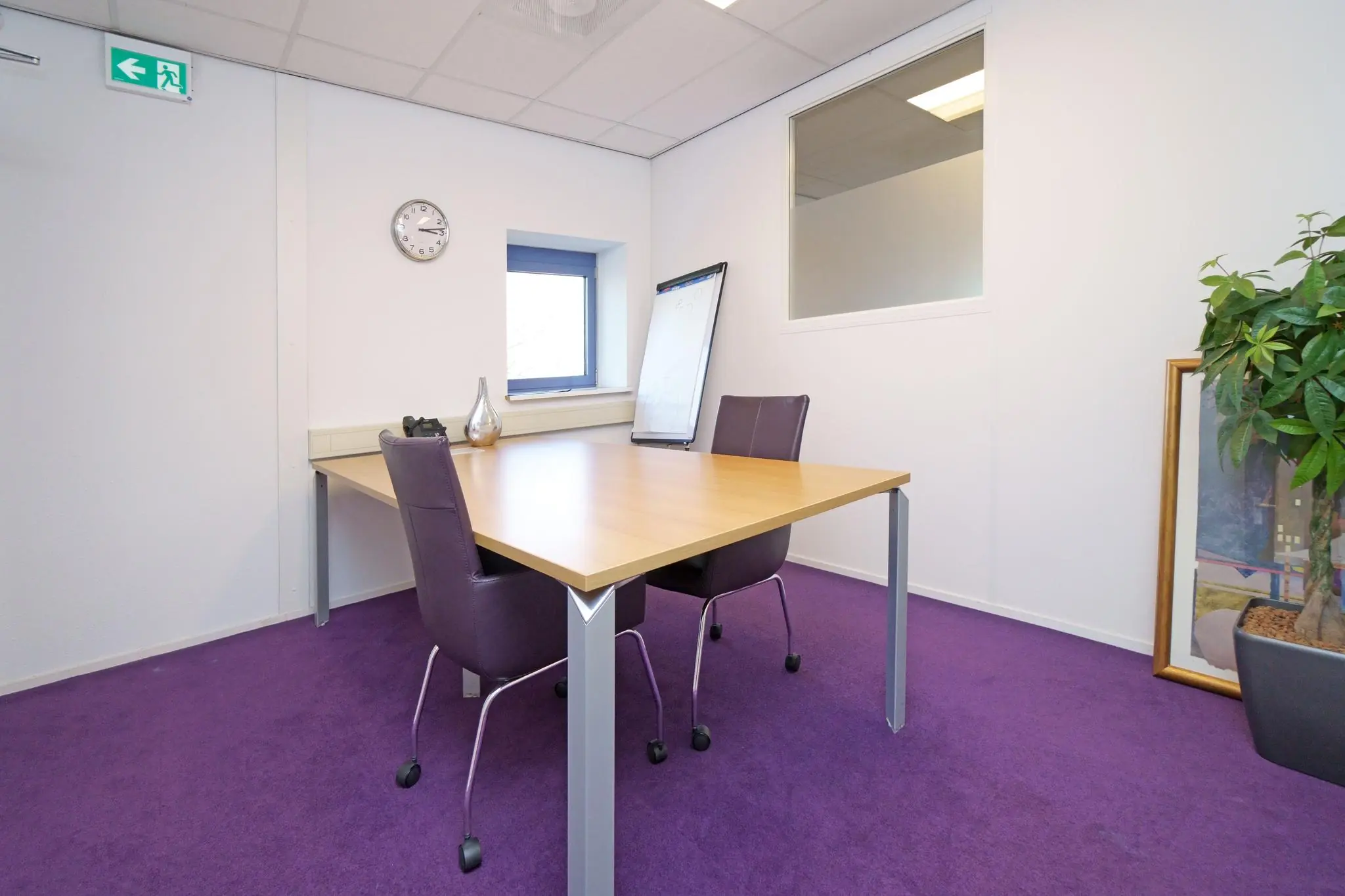 Kleine vergaderruimte met paarse vloerbedekking, twee paarse stoelen, een houten tafel en een whiteboard, gelegen aan de Zuiderweg.