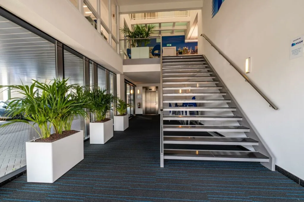 Moderne entreehal aan de Bahialaan met metalen trap, grote ramen en groene planten in witte bakken.
