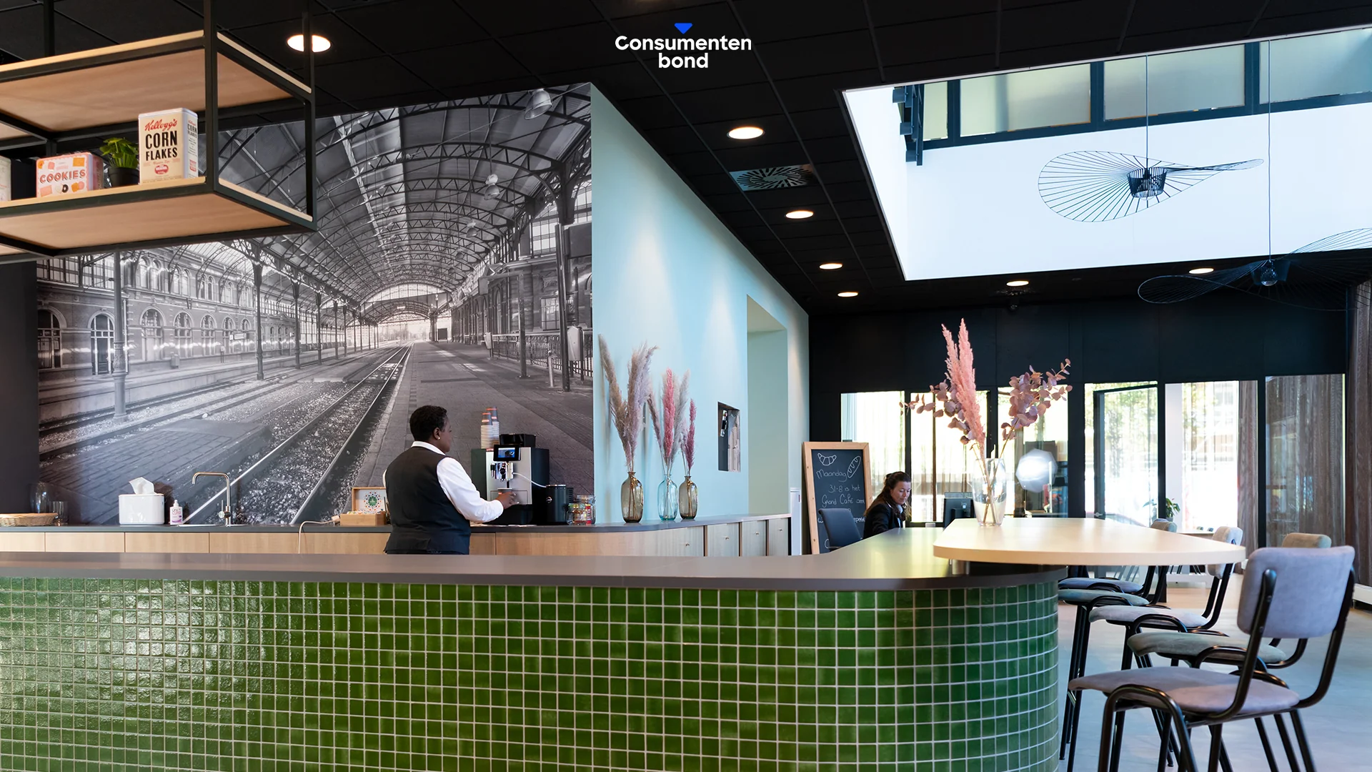Moderne ontvangstruimte van de Consumentenbond aan het Enthovenplein met groene tegelbalie, koffieautomaat en muurdecoratie van een historisch treinstation.