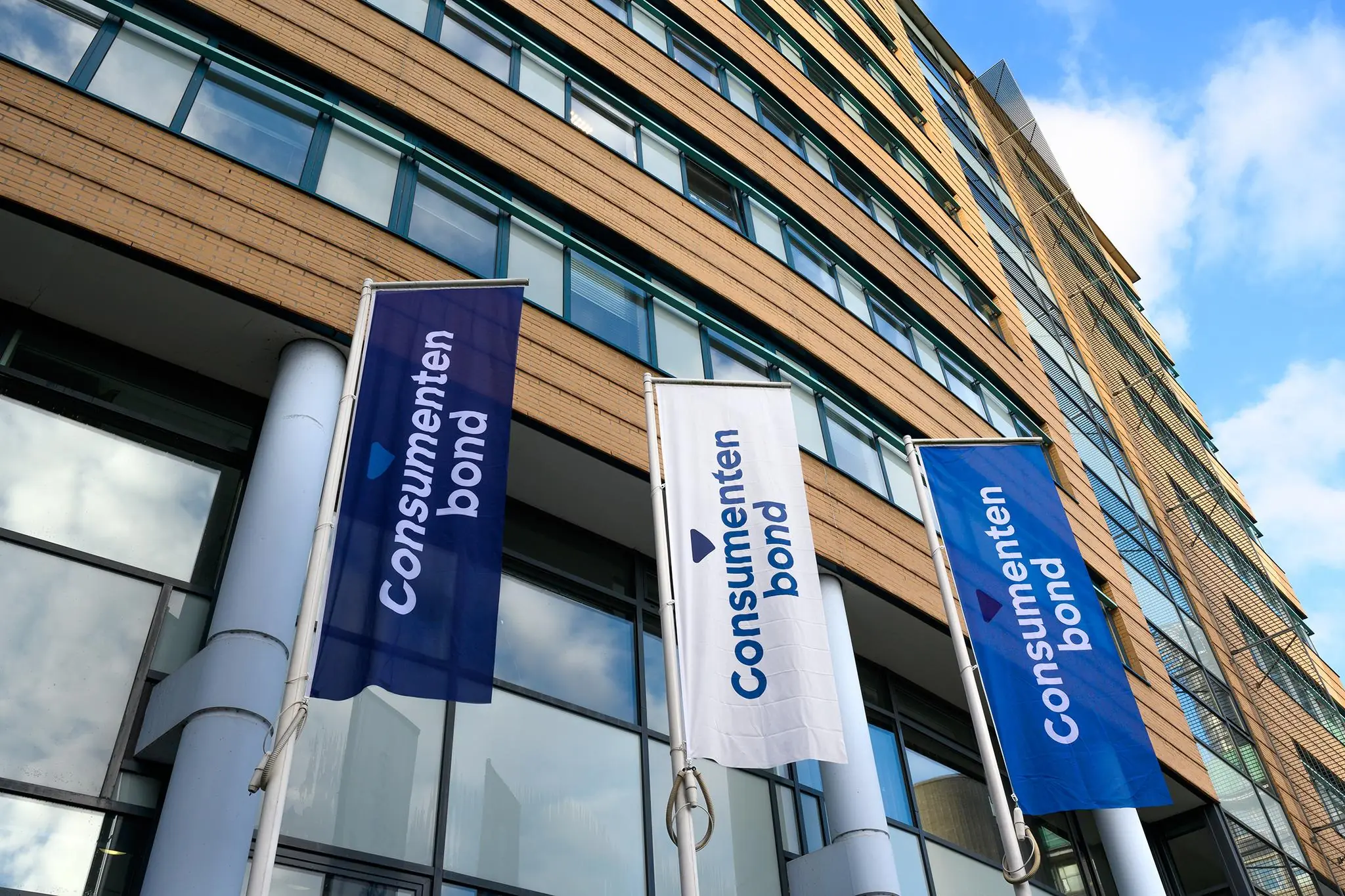 Vlaggen met het logo van de Consumentenbond wapperen voor een modern kantoorgebouw aan het Enthovenplein.
