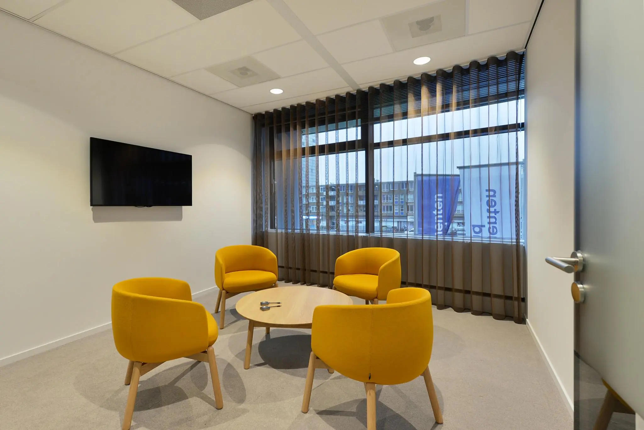 Vergaderruimte aan het Enthovenplein met vier gele stoelen, een ronde tafel en een flatscreen-tv aan de muur.