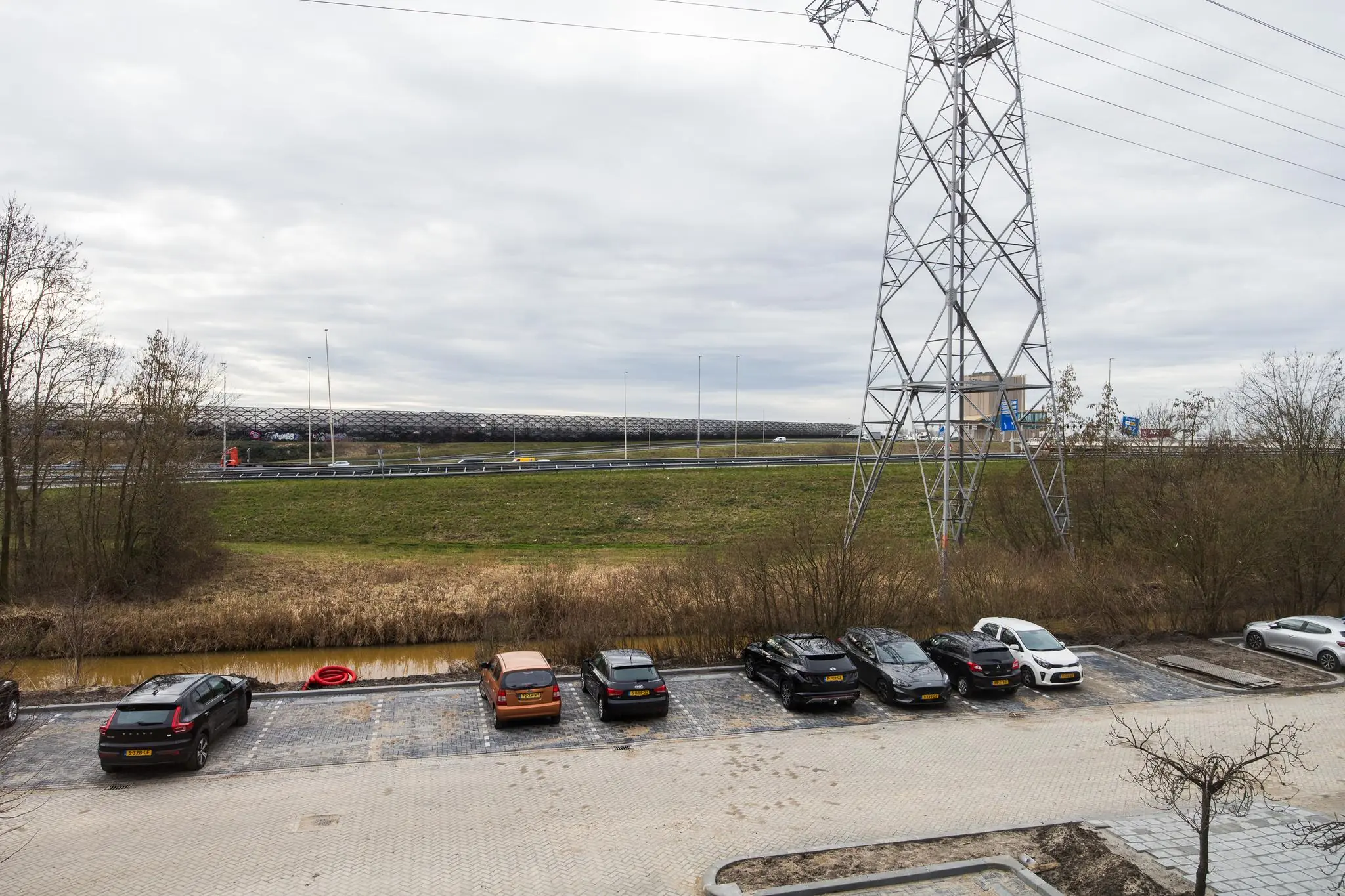 Parkeerplaats aan de Zonnebaan met geparkeerde auto's, een hoogspanningsmast en zicht op de snelweg met geluidsscherm.