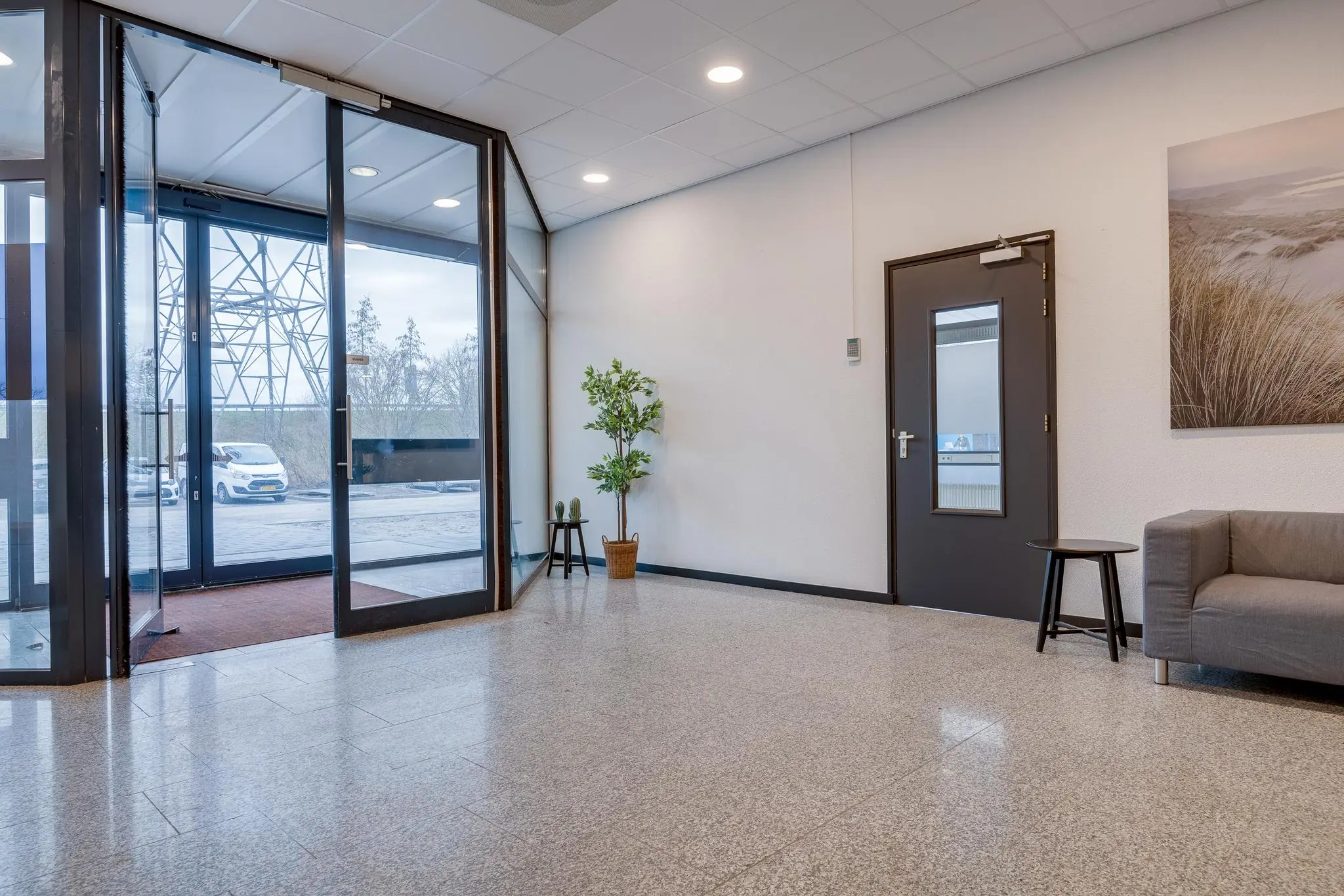 Ingang en wachtruimte van een modern kantoorpand aan de Zonnebaan met glazen deuren, een plant en een grijze bank.
