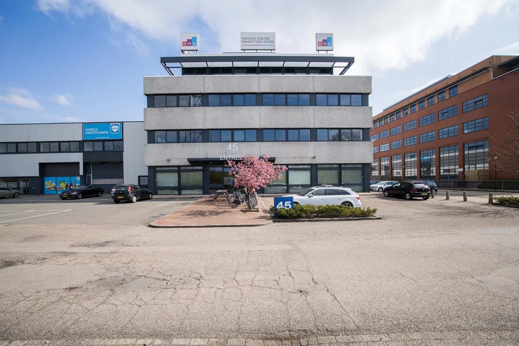 Kantoorpand aan de Zonnebaan 45 met parkeerplaatsen, bloeiende boom en omliggende bedrijfsgebouwen.