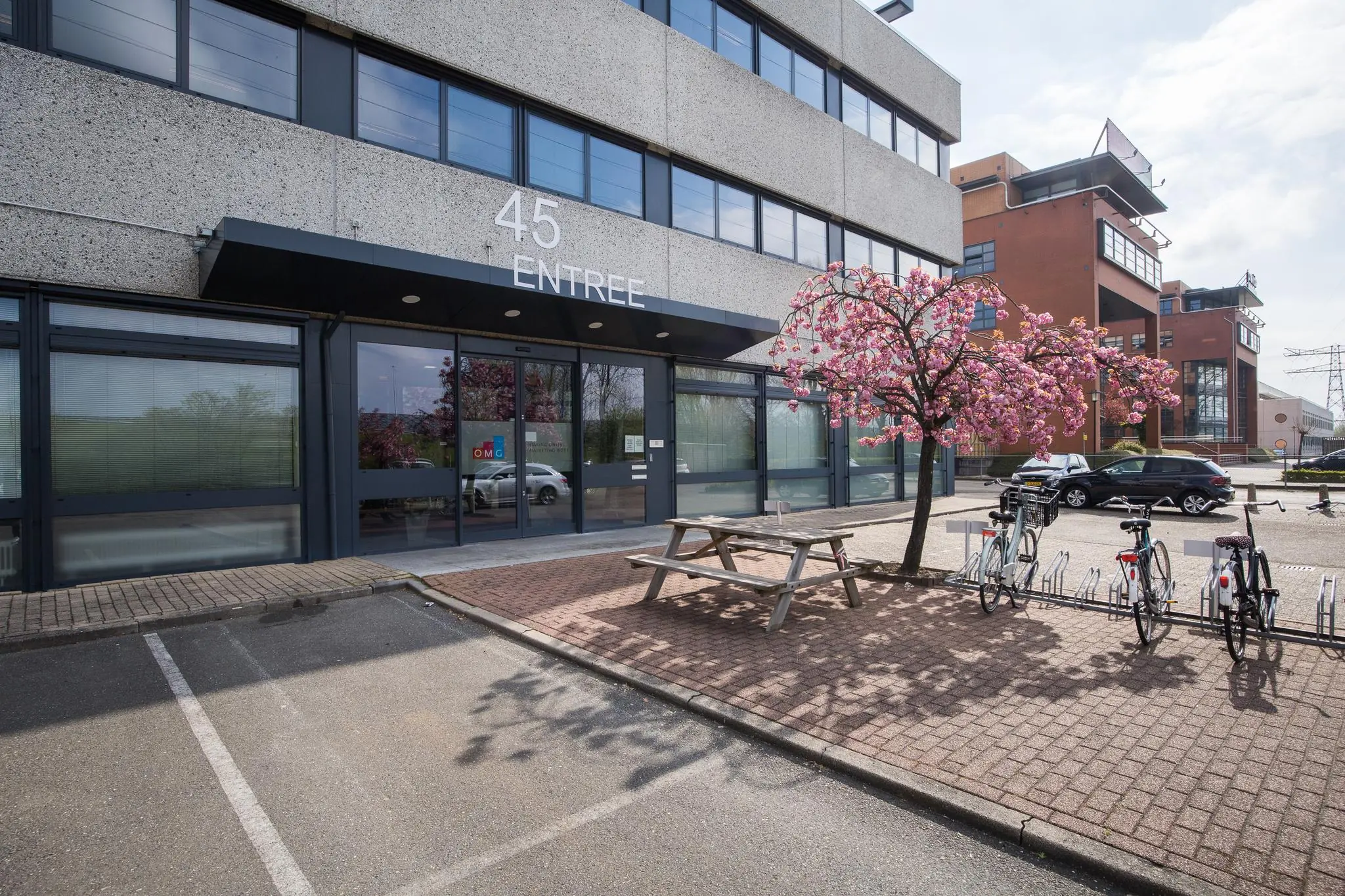 Ingang van een kantoorgebouw aan de Zonnebaan 45 met fietsenrekken, picknicktafel en een bloeiende boom op de stoep.