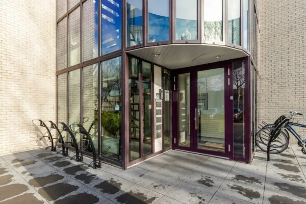 Ingang van een modern appartementencomplex aan de Plesmanstraat met glazen gevel, paarse deuren en fietsenrekken.