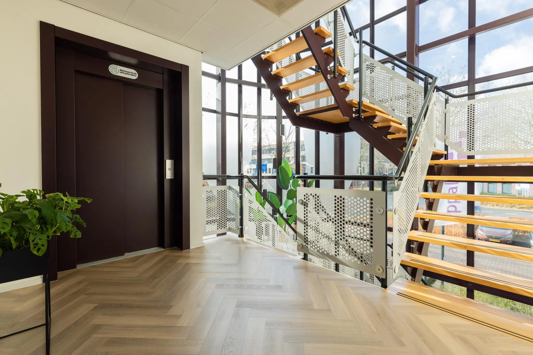Trapportaal met houten trap, lift en grote ramen in een modern kantoorgebouw aan de Plesmanstraat.
