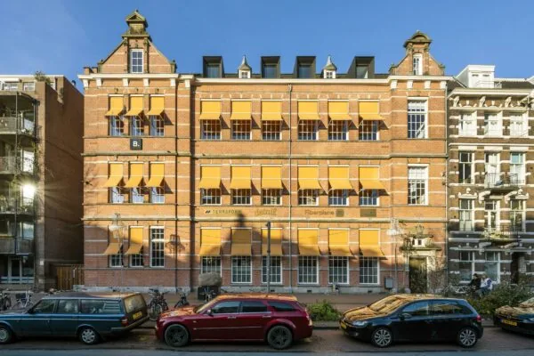 Gevelaanzicht van kantoorgebouw B. aan de Overtoom met rode bakstenen, gele zonneschermen en geparkeerde auto's.