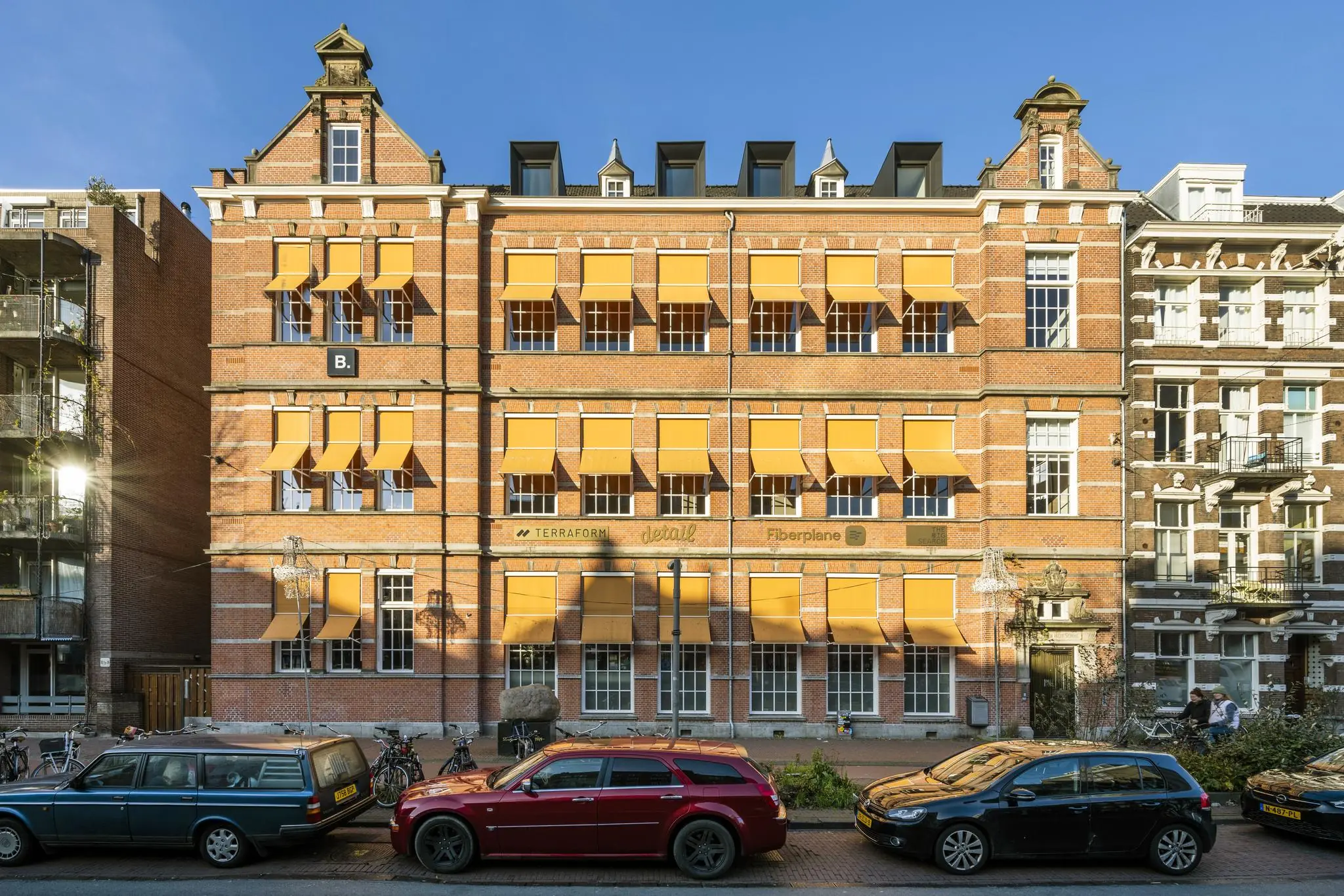Gevelaanzicht van kantoorgebouw B. aan de Overtoom met rode bakstenen, gele zonneschermen en geparkeerde auto's.