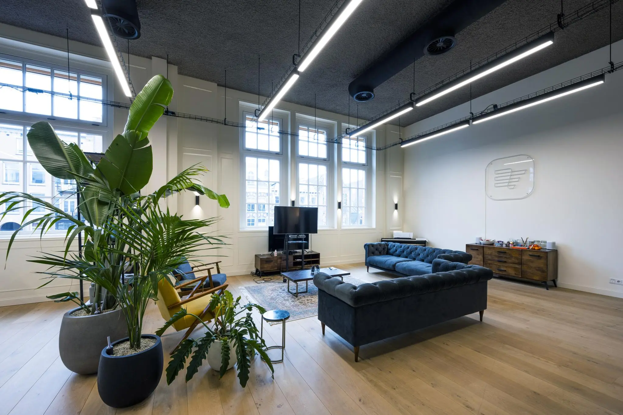 Modern ingerichte lounge- of wachtruimte aan de Overtoom met grote ramen, planten, blauwe banken en een flatscreen-tv.