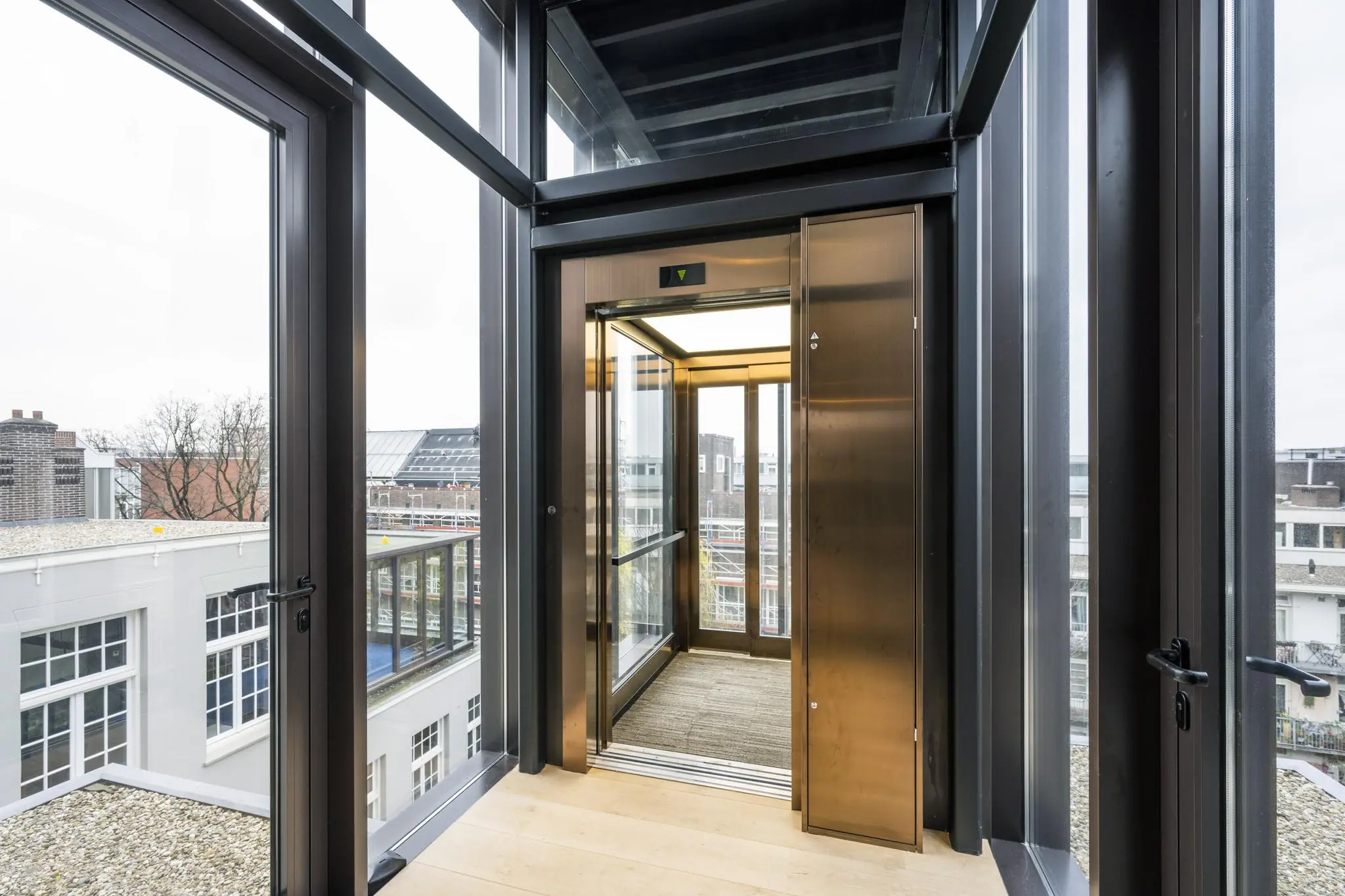 Moderne glazen lift met bronzen deuren in een transparante liftschacht met uitzicht op daken aan de Overtoom.