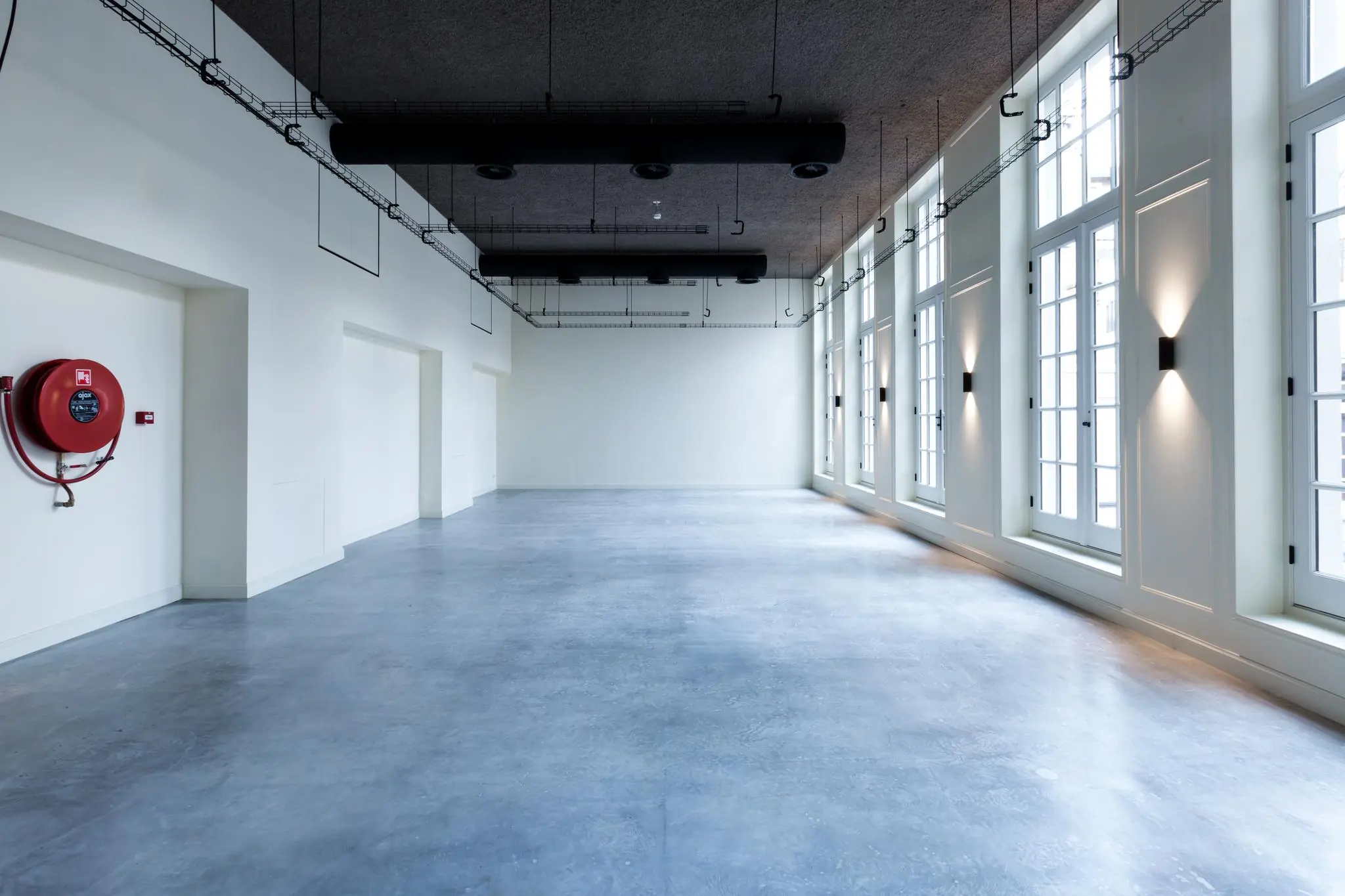 Lichte, lege ruimte met grote ramen, een betonnen vloer en een brandslanghaspel aan de muur in gebouw Overtoom.