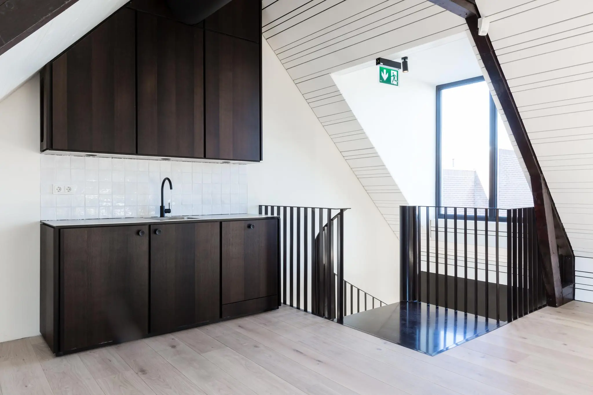 Moderne zolderruimte met een donkerhouten kitchenette, witte tegelachterwand en zwart metalen trapleuning bij een groot raam.