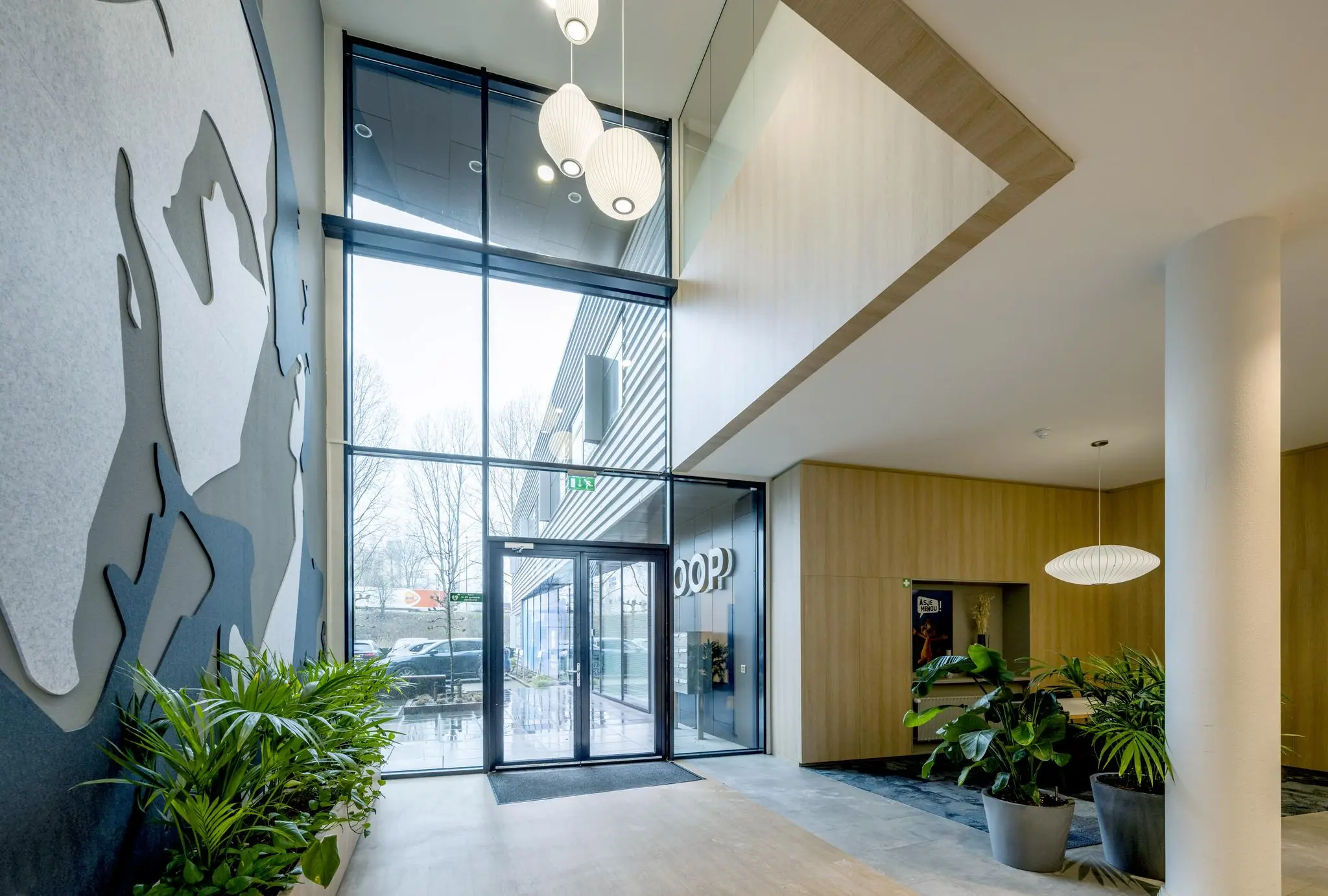 Moderne entreehal aan de Joop Geesinkweg met hoge glazen deuren, hanglampen, kunst aan de muur en veel groen.
