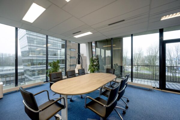 Vergaderruimte met ovale tafel en acht stoelen in een modern kantoorpand aan de Joop Geesinkweg, met uitzicht op omliggende gebouwen en bomen.