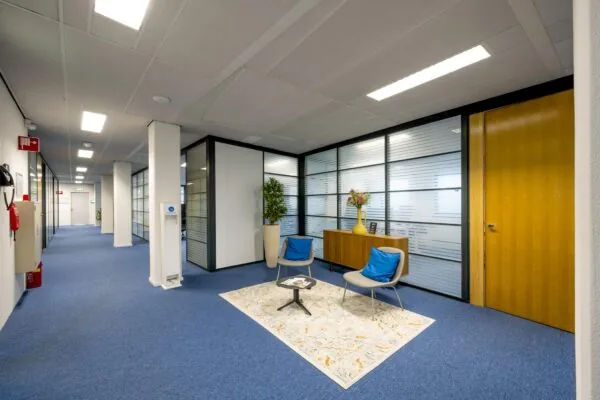 Moderne kantoorhal aan de Joop Geesinkweg met glazen scheidingswanden, blauwe vloerbedekking en een wachthoek met twee stoelen.