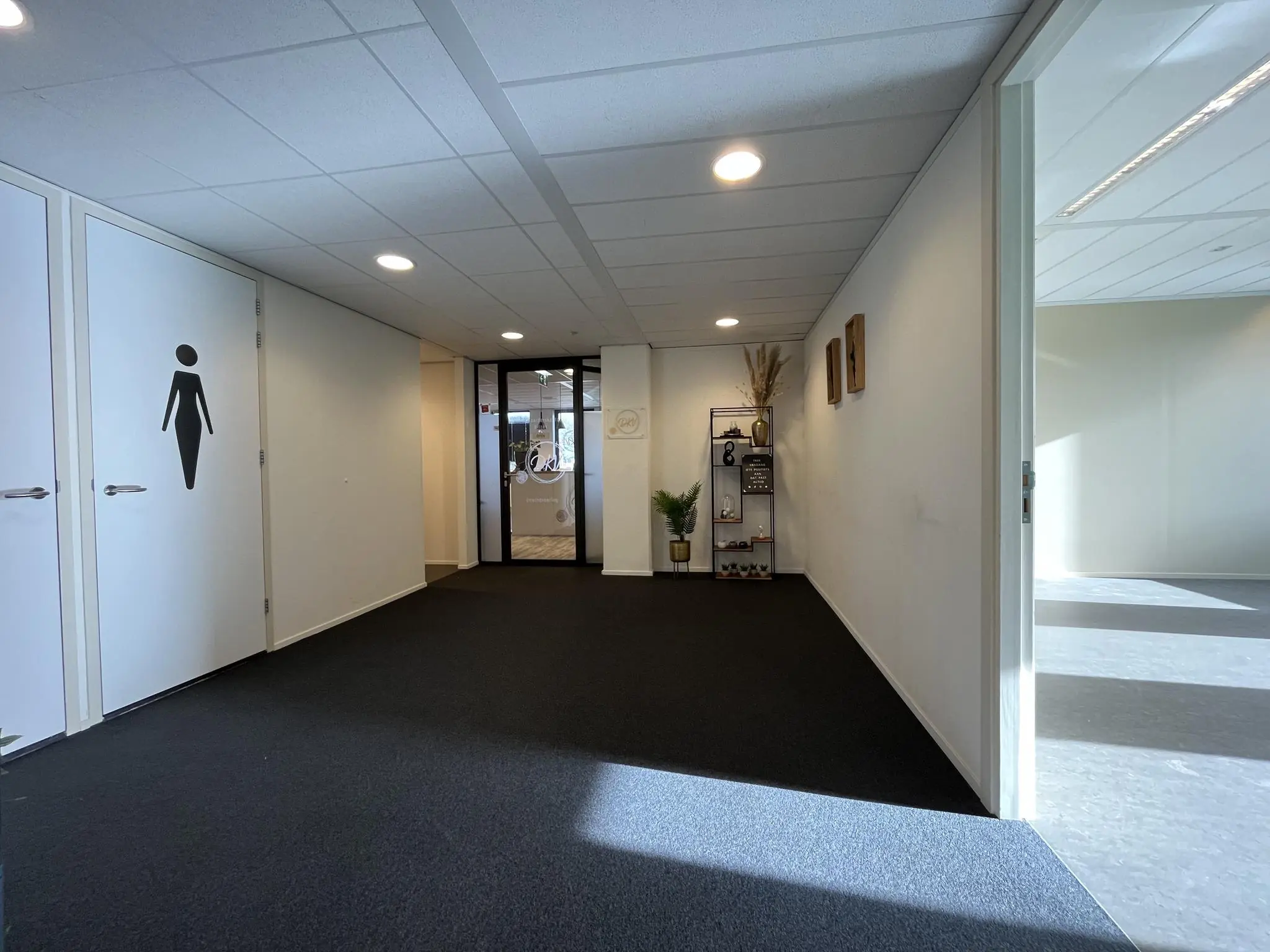 Verlichte gang in een kantoorpand met een damestoiletdeur, decoratieve plank en toegang tot de ruimte van Praktijk de Ratelaar.