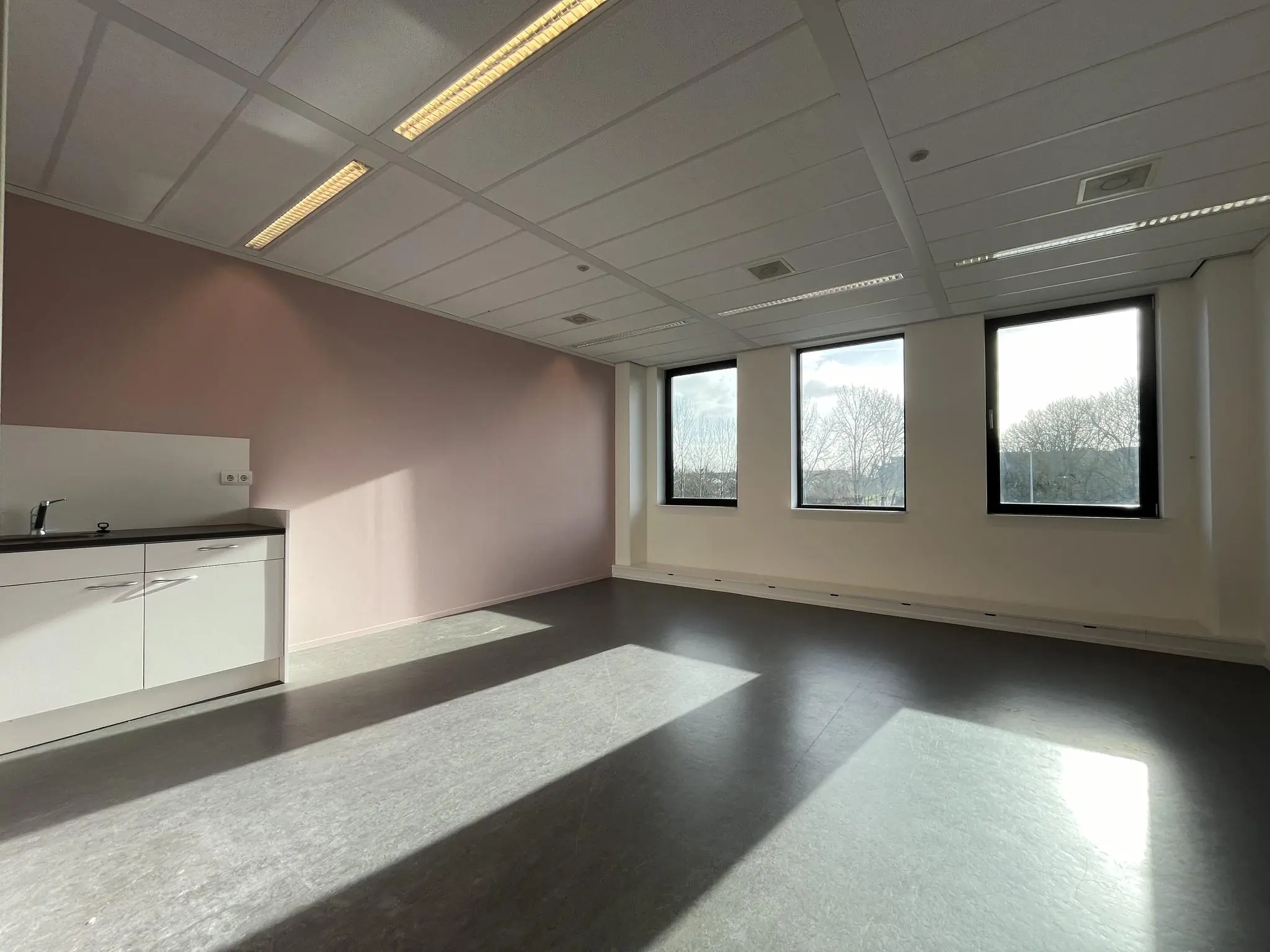 Lichte, lege kantoorruimte met drie grote ramen, een roze muur en een wit keukenblok aan de linkerkant.