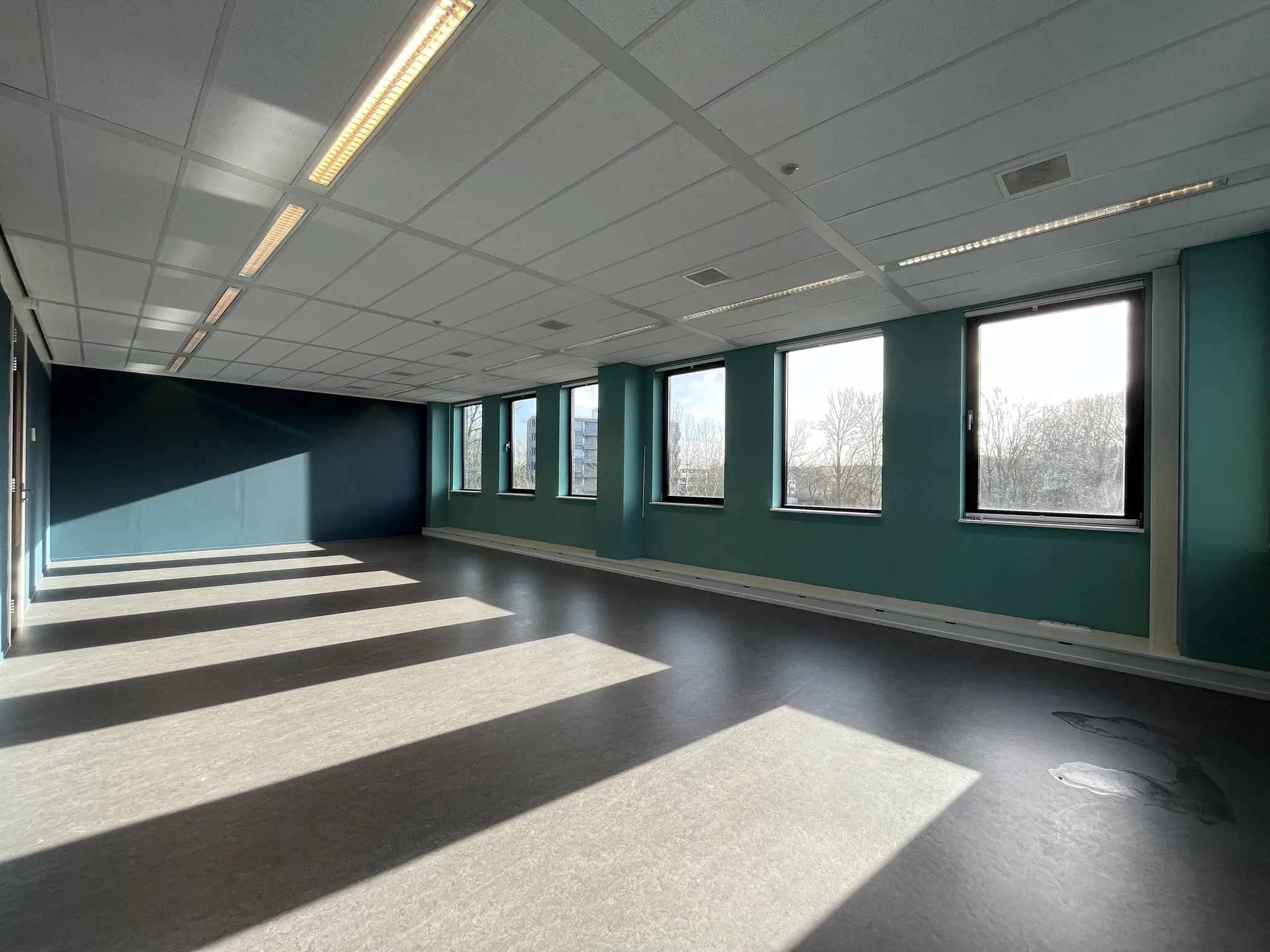 Lege kantoorruimte in gebouw Ratelaar met grote ramen en zonlicht dat schaduwen werpt op de vloer.