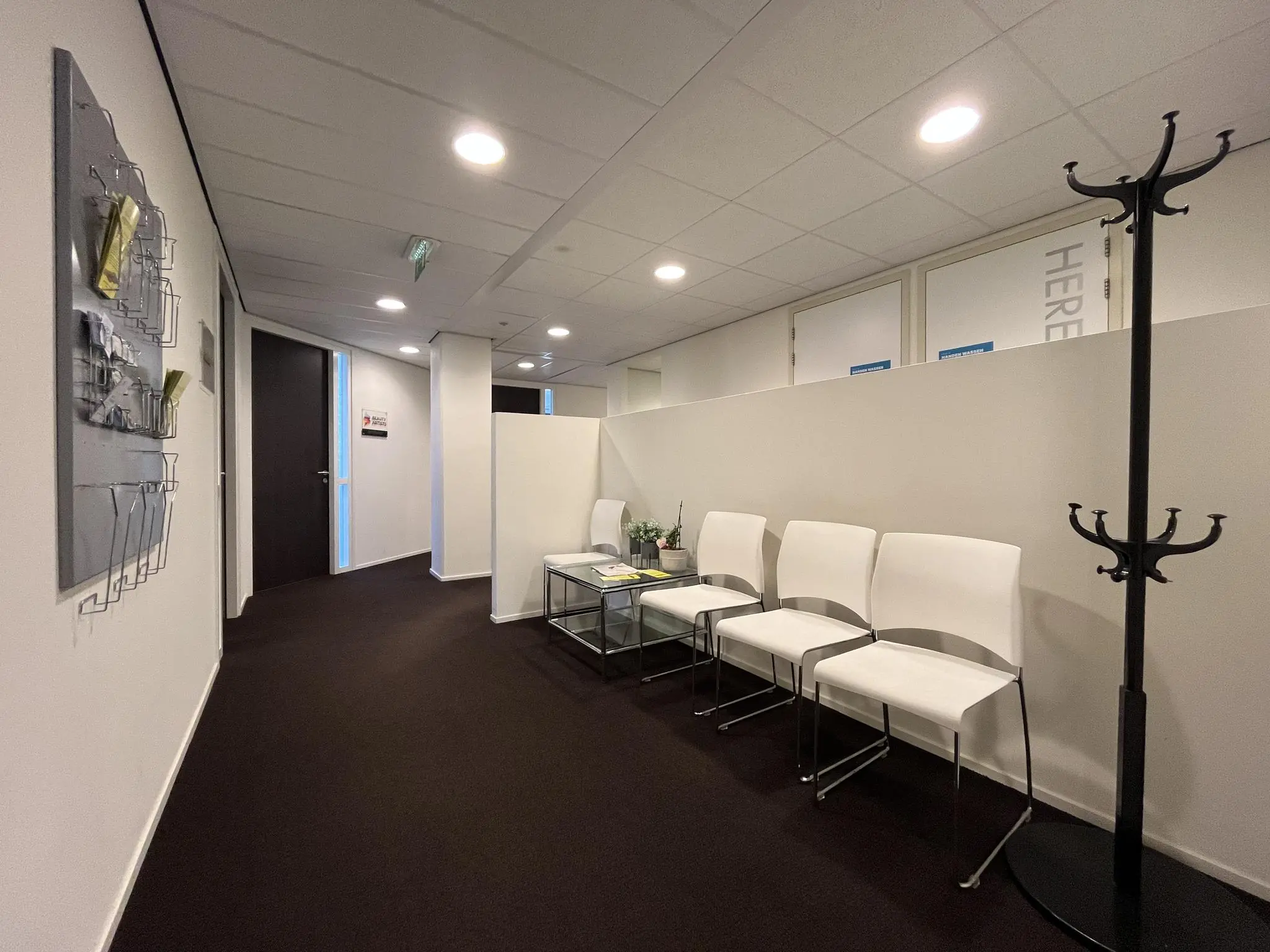 Wachtruimte met vier witte stoelen, een glazen bijzettafel en een kapstok in een modern kantoorpand.