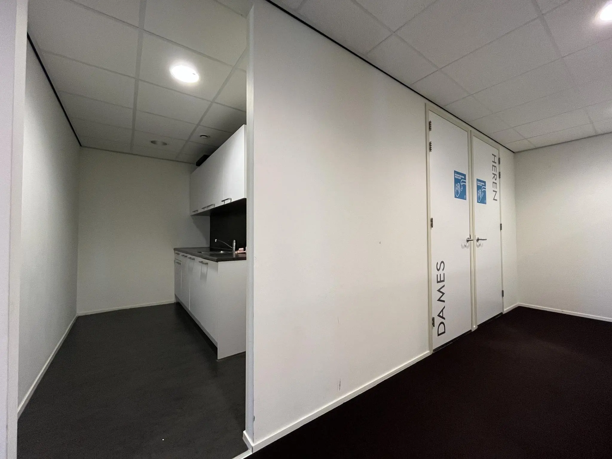 Witte gang met een kleine keukenhoek en twee toiletten aangeduid als dames en heren.