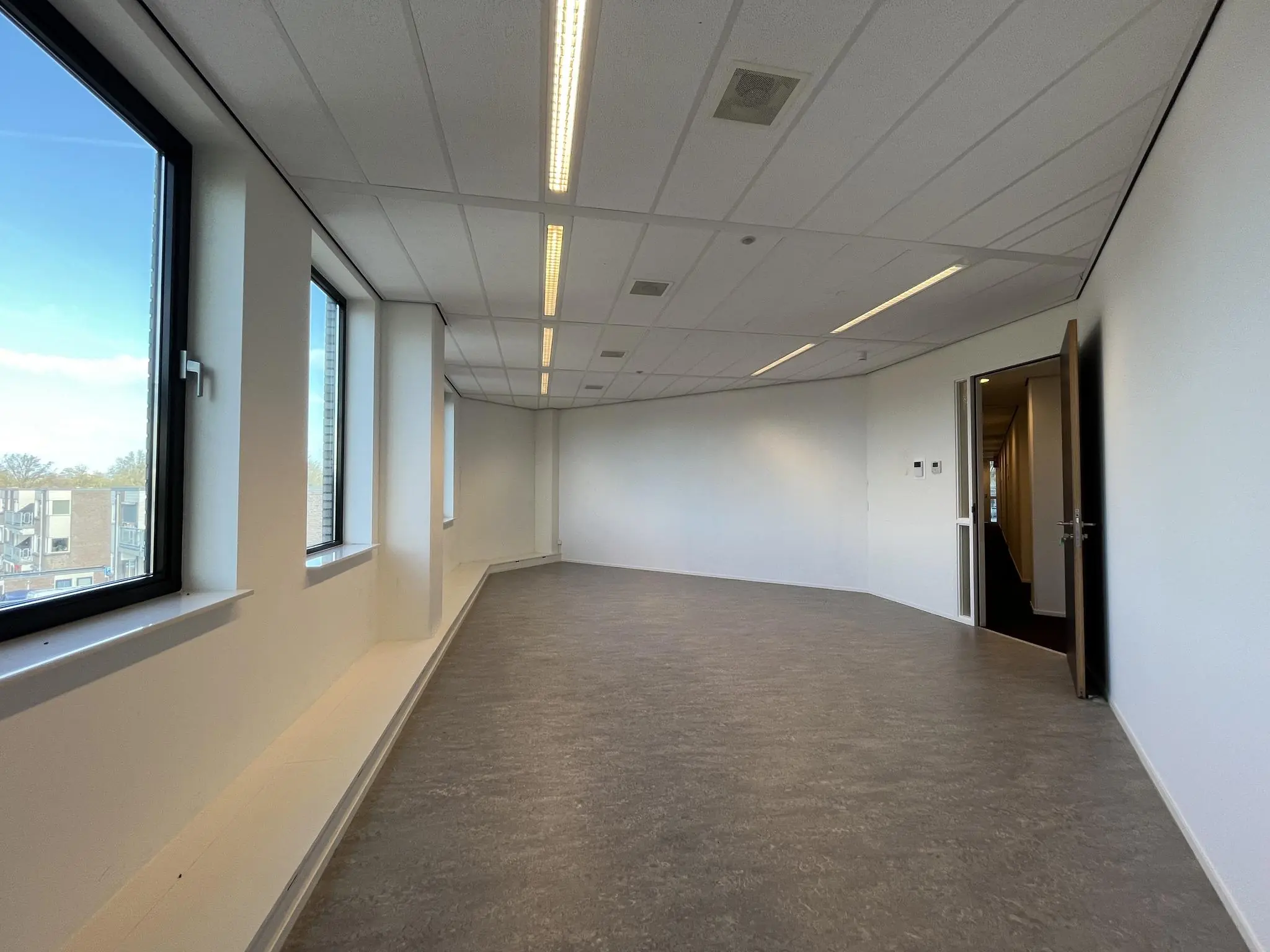 Leeg kantoorruimte met grote ramen, witte muren en een open deur naar een gang in het gebouw Ratelaar.