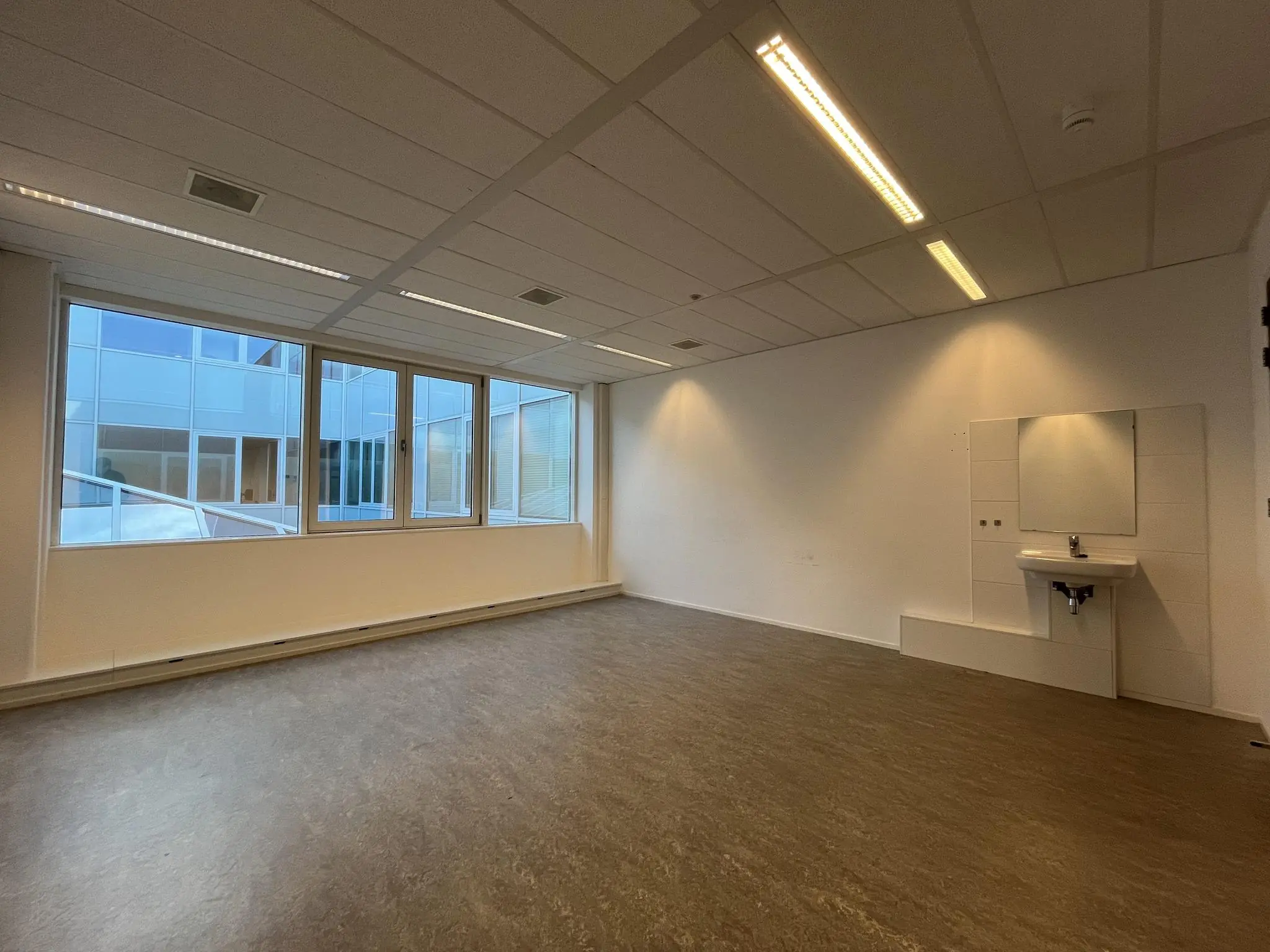 Lege ruimte met een wastafel en grote ramen met uitzicht op een binnenplaats van een modern gebouw.