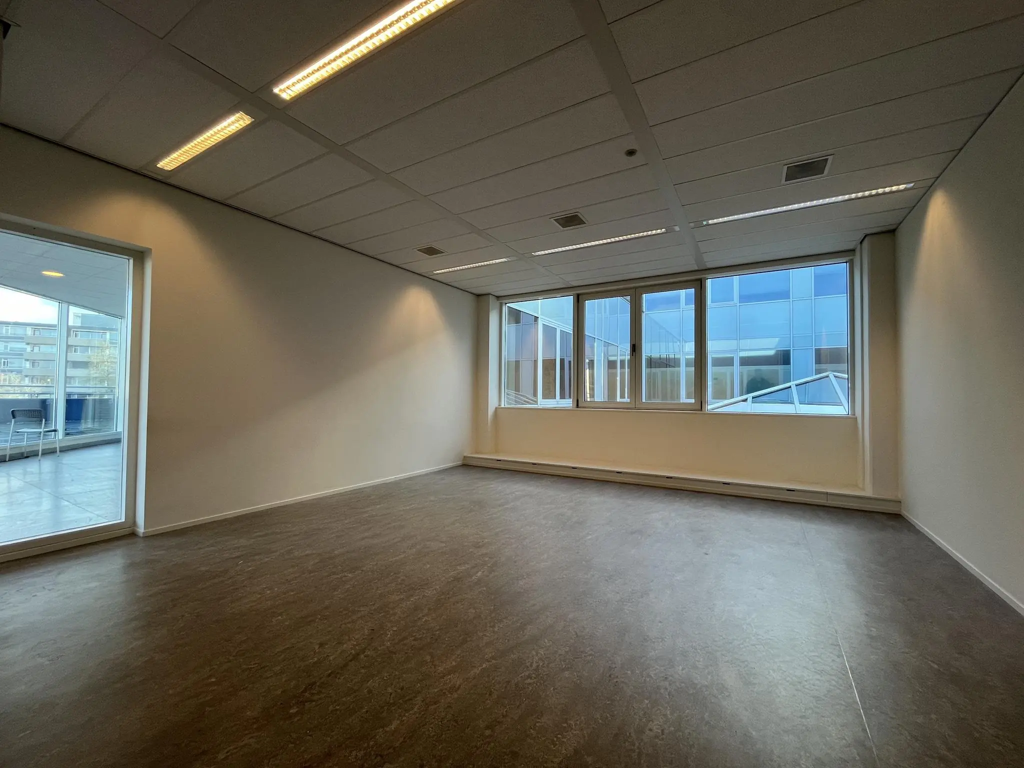 Lege kantoorruimte met grote ramen en uitzicht op een modern kantoorgebouw.