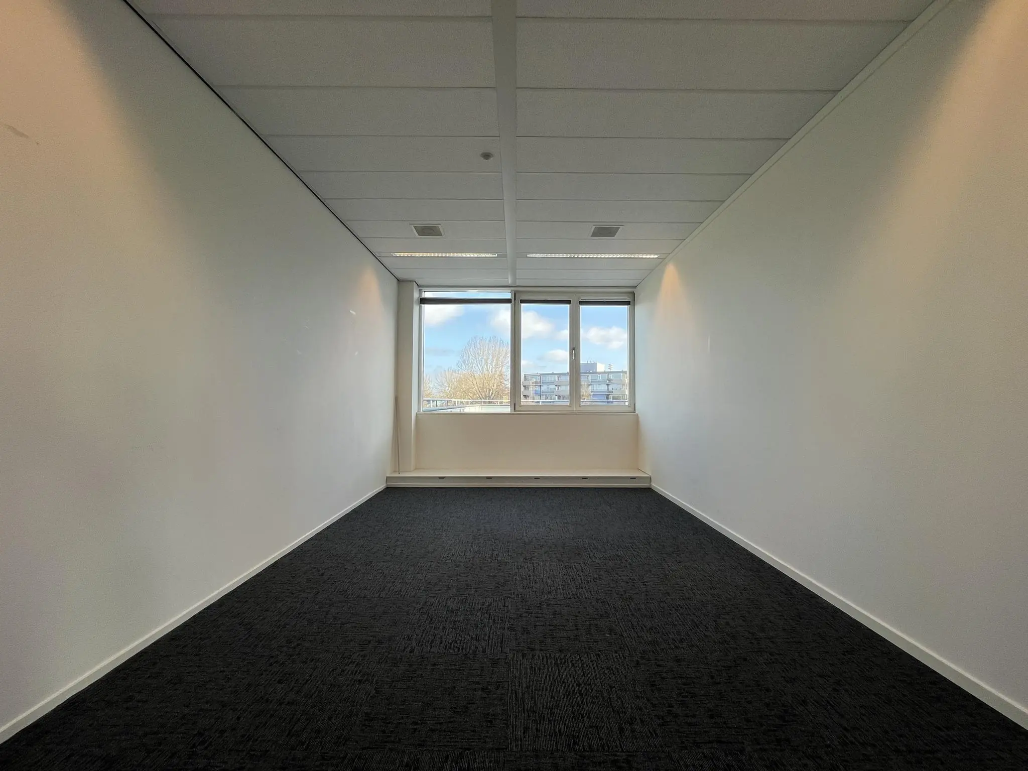 Lege, smalle kantoorruimte met witte muren, donkergrijze vloerbedekking en een raam met uitzicht op gebouwen en bomen.