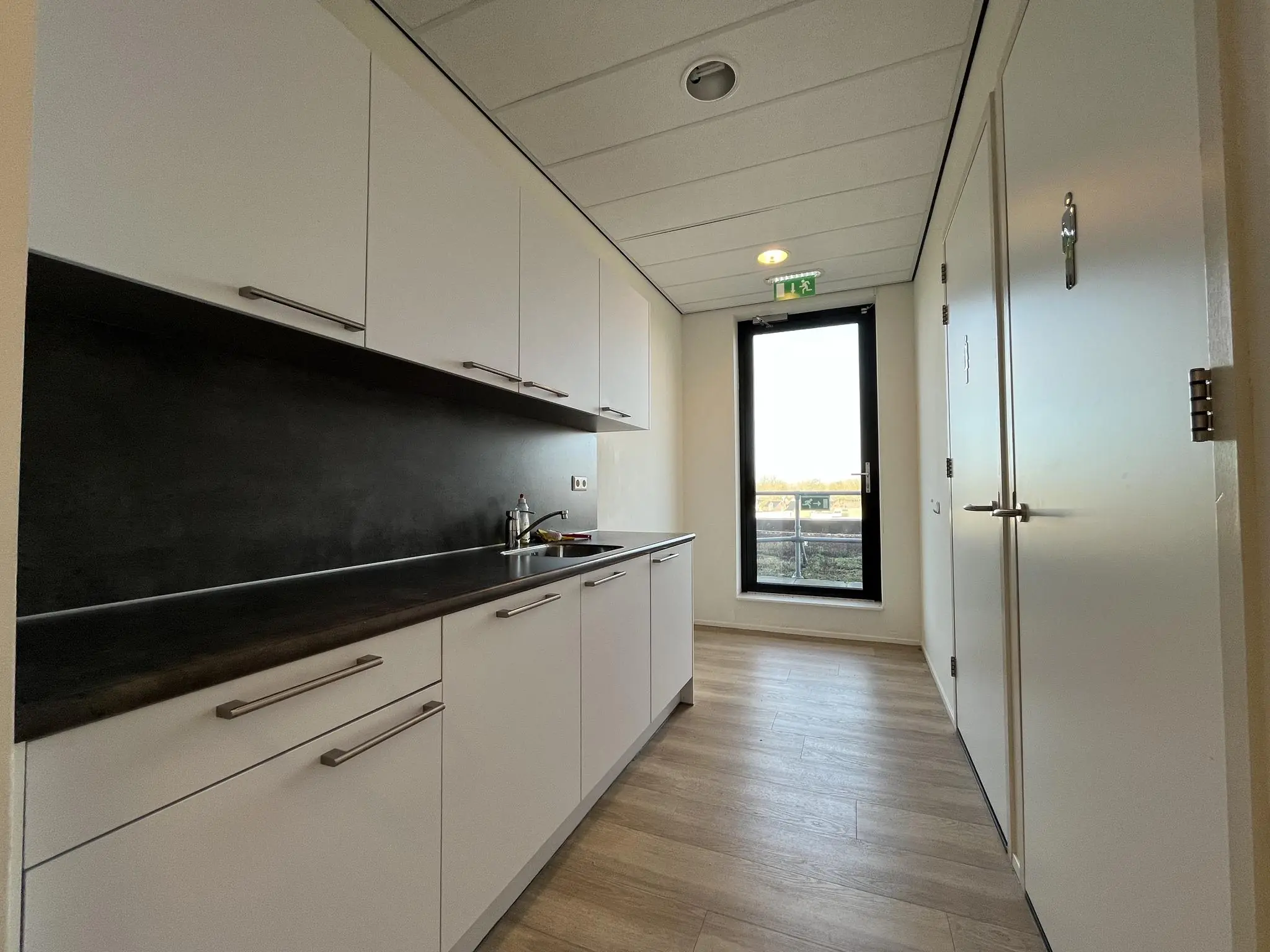 Moderne pantry met witte keukenkasten, donker aanrechtblad en een glazen deur met uitzicht naar buiten.