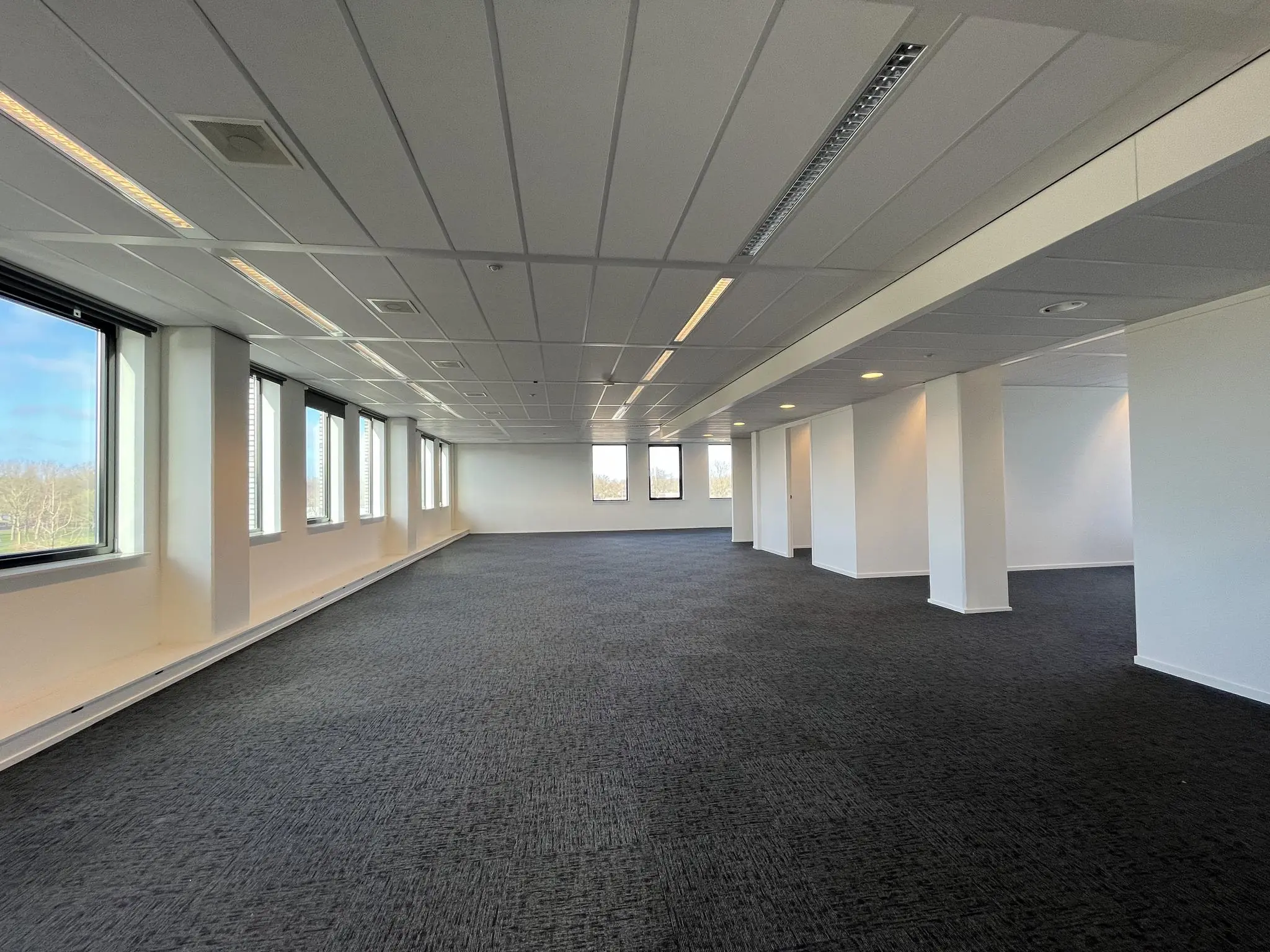 Ruime, lege kantoorruimte in gebouw Ratelaar met donkere vloerbedekking en grote ramen langs één zijde.