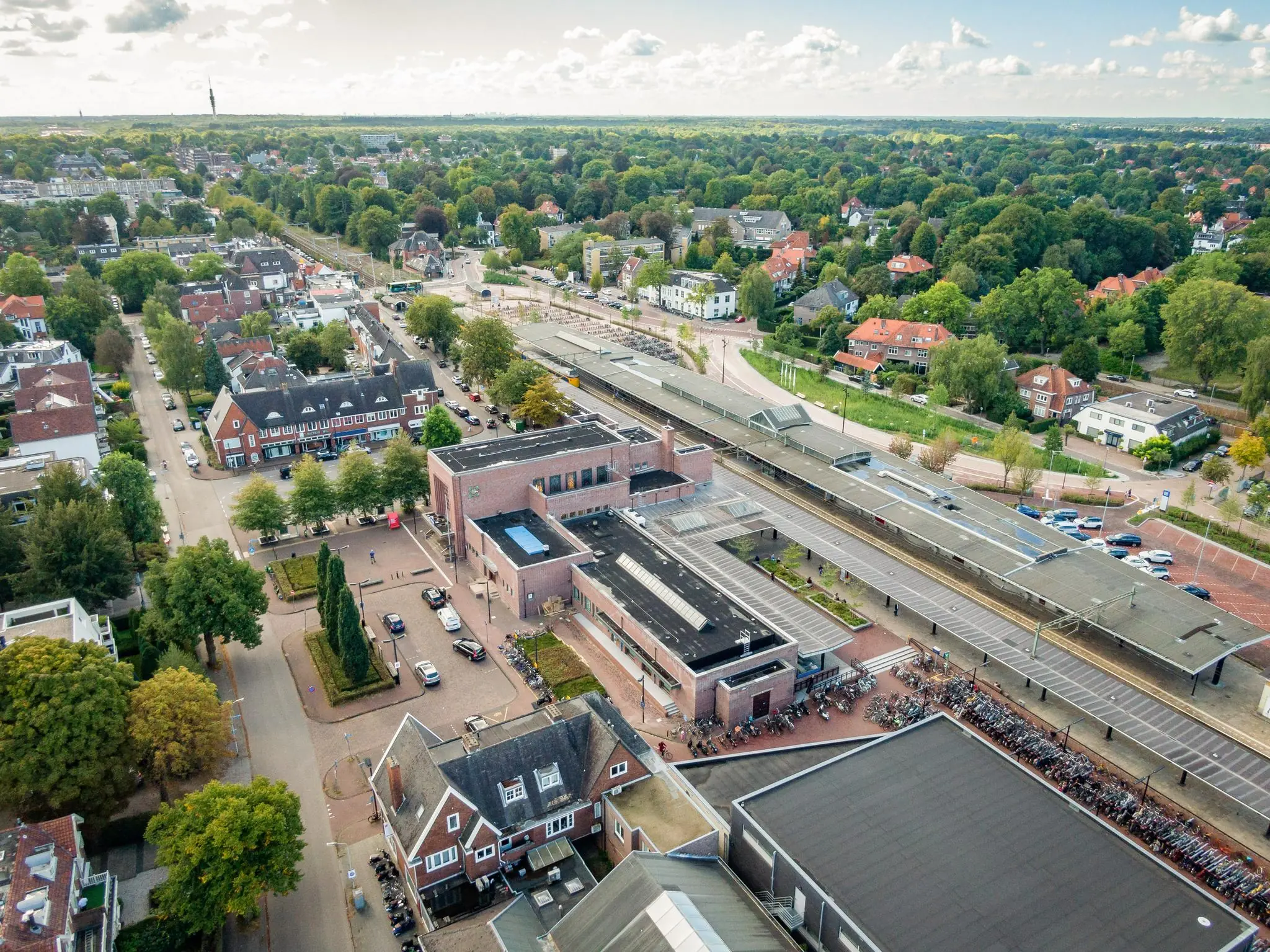 Luchtfoto van het Stationsplein in een groene woonwijk met het treinstation, fietsenstallingen en omliggende bebouwing.