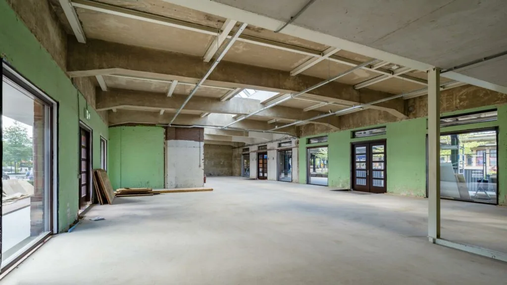 Leegstaand en deels gerenoveerd winkel- of kantoorpand aan het Stationsplein met zicht op grote ramen en betonnen plafond.
