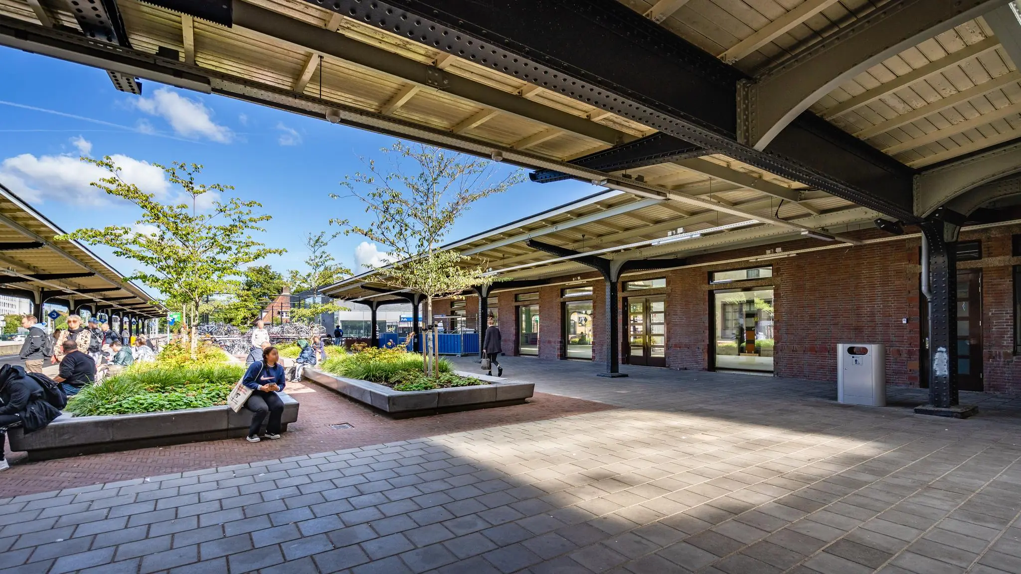 Overdekt stationsplein met zitmogelijkheden, groenstroken en wachtende reizigers op een zonnige dag.