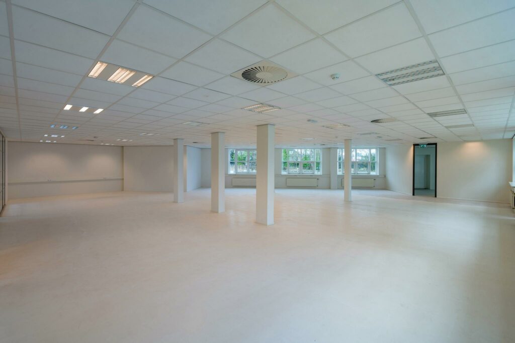 Lichte, lege kantoorruimte met witte muren, systeemplafond en ramen aan één zijde in het Rhijnspoorgebouw.