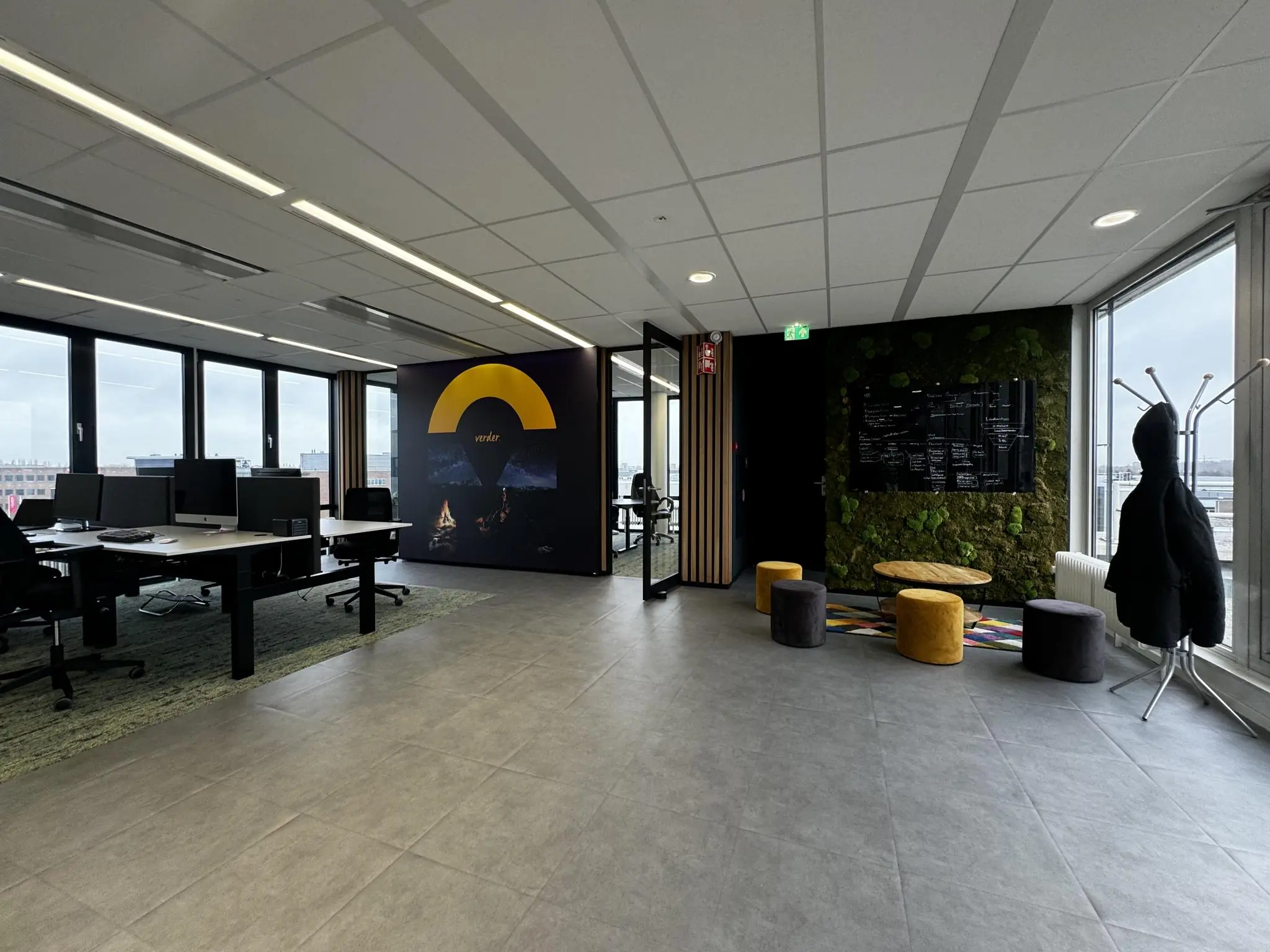 Moderne kantoorruimte in Rijnzathe met werkplekken, een moswand, zitpoefjes en een zwarte jas aan een kapstok.