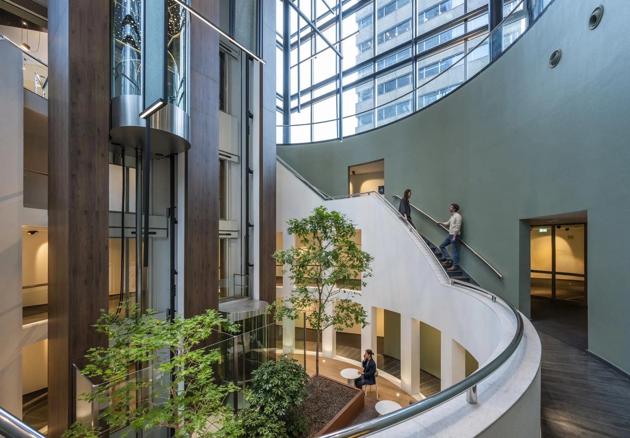 Modern atrium in Amstelplein met glazen liften, binnentuin en mensen op een trap en zitplaatsen.