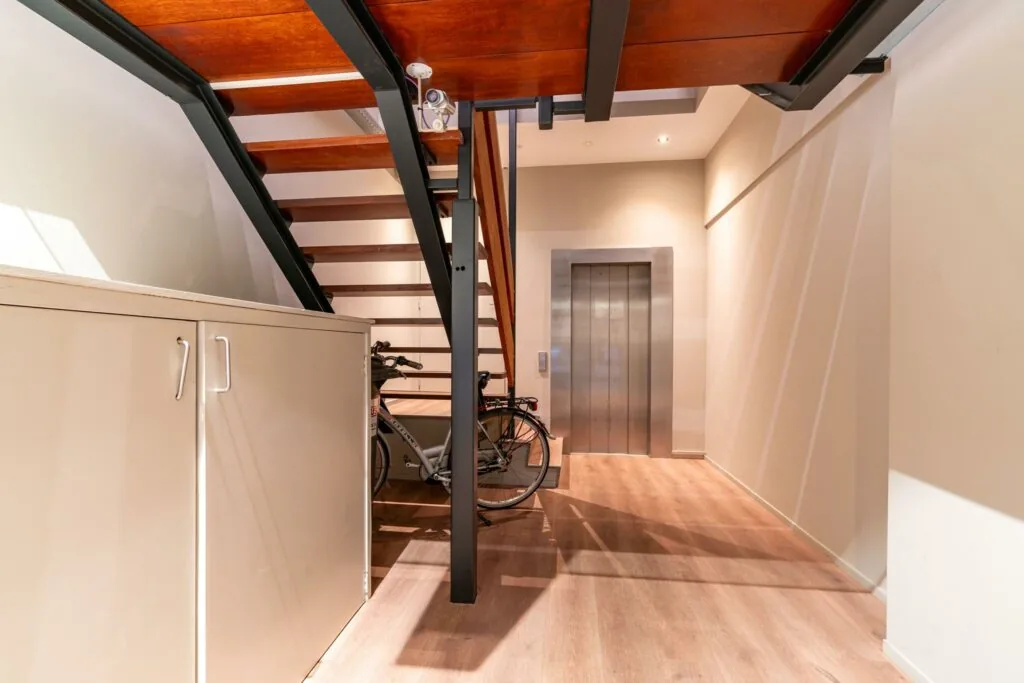 Moderne hal met houten trap, liftdeur, fiets en opbergkast op een lichte houten vloer.