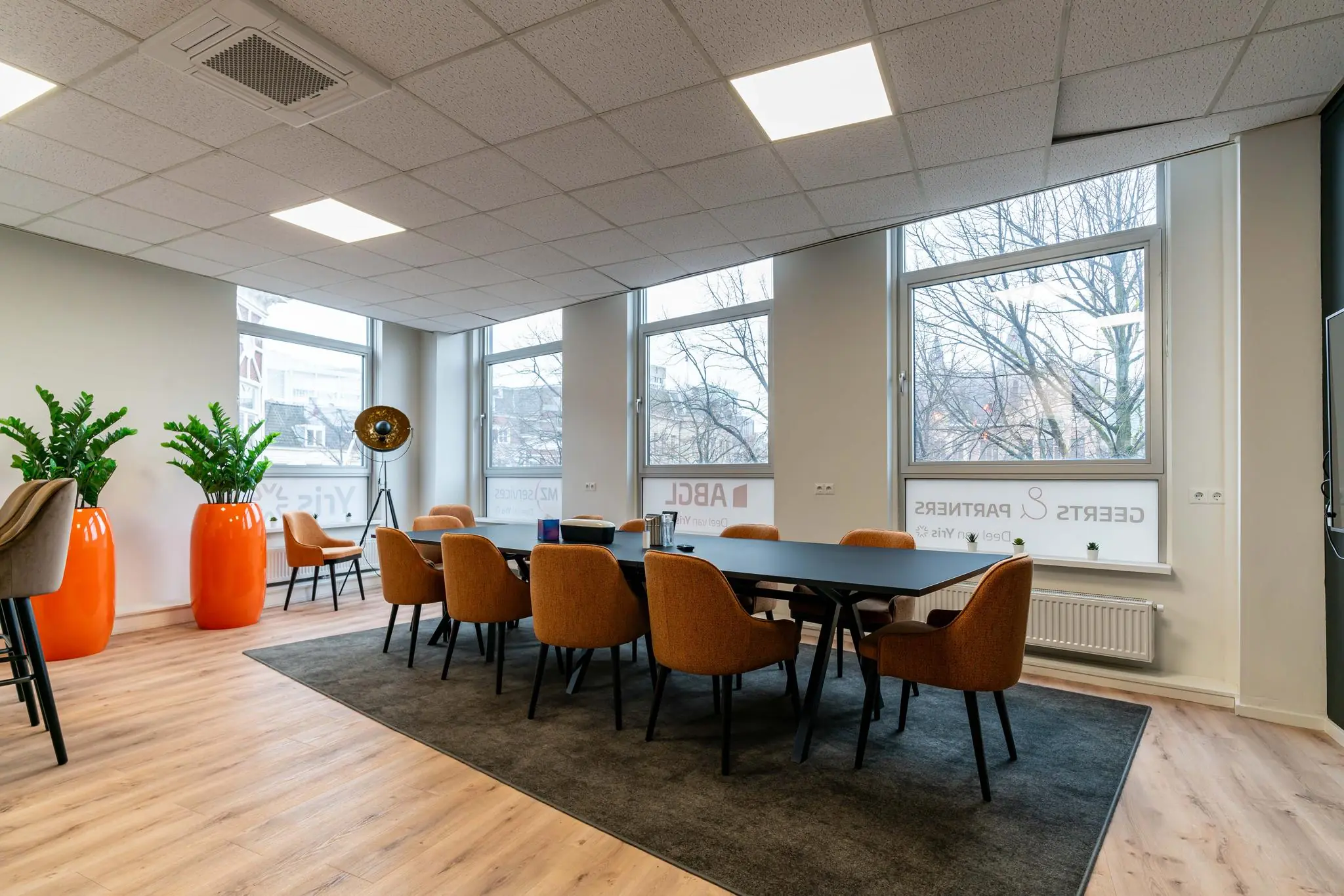 Moderne vergaderruimte met grote ramen, oranje stoelen en opvallende oranje plantenpotten.