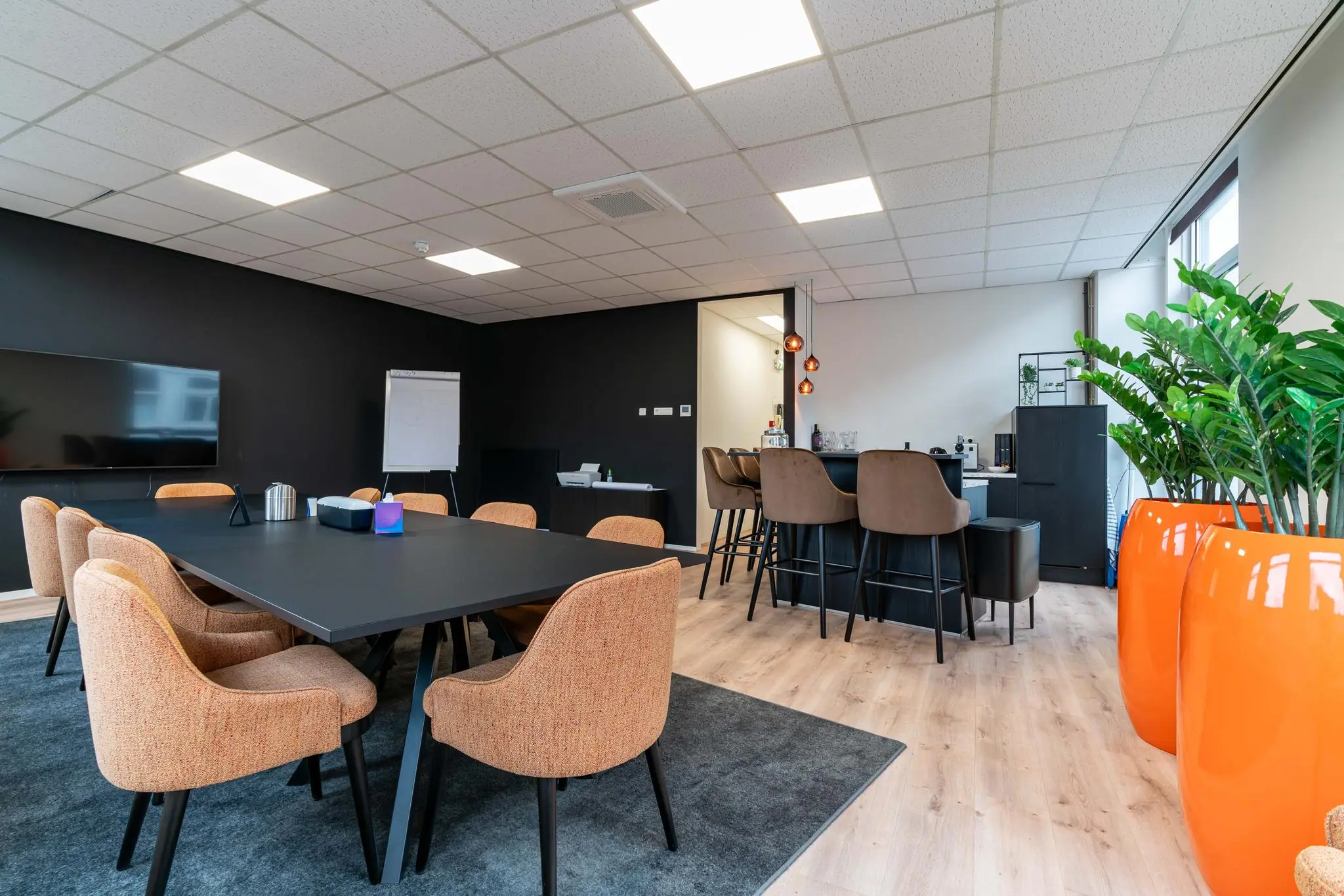 Moderne vergaderruimte met een grote tafel, oranje stoelen, barhoek en opvallende oranje plantenbakken.