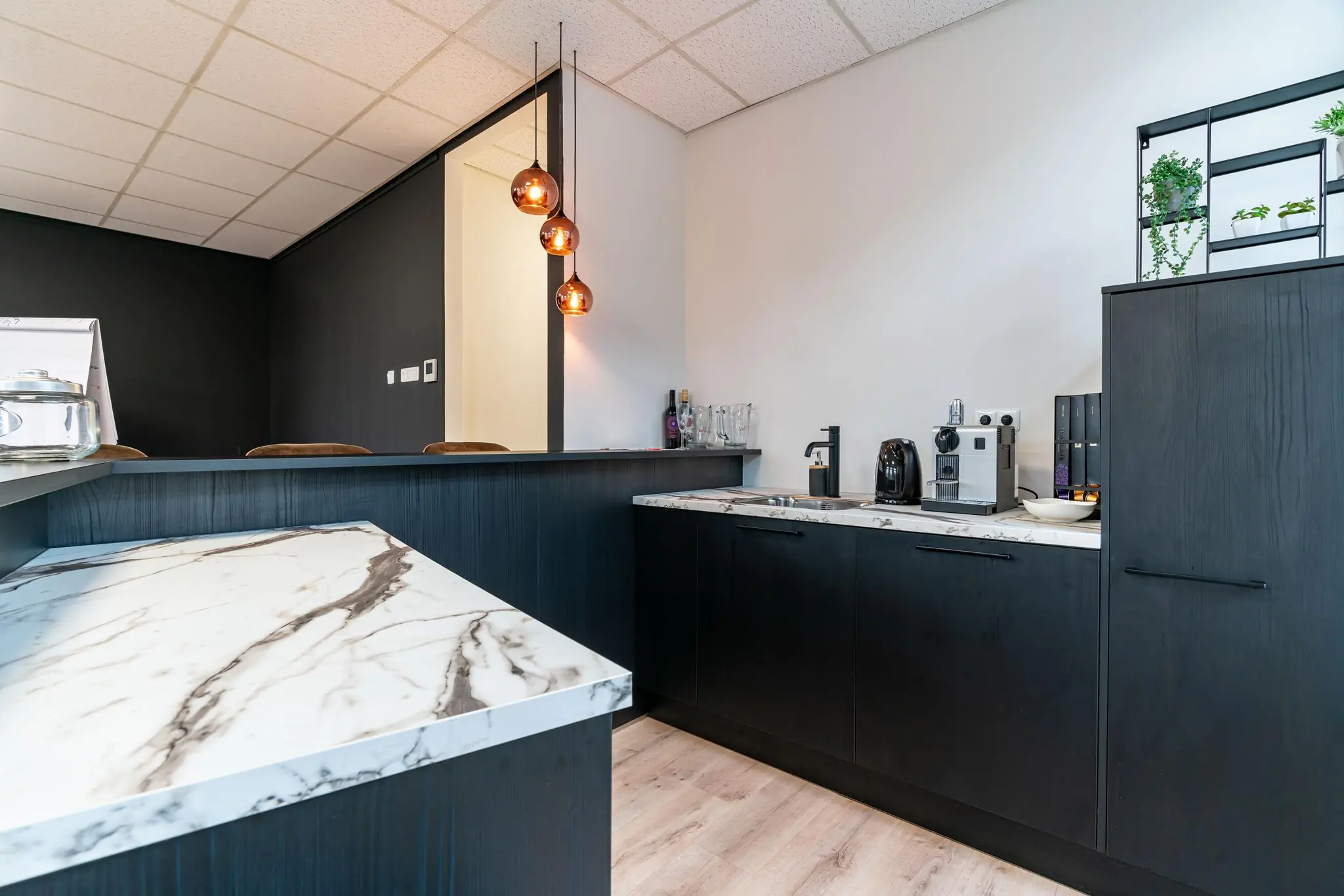 Moderne kantoorkeuken met zwart-witte marmeren werkbladen, koffieapparaten en hangende glazen lampen.