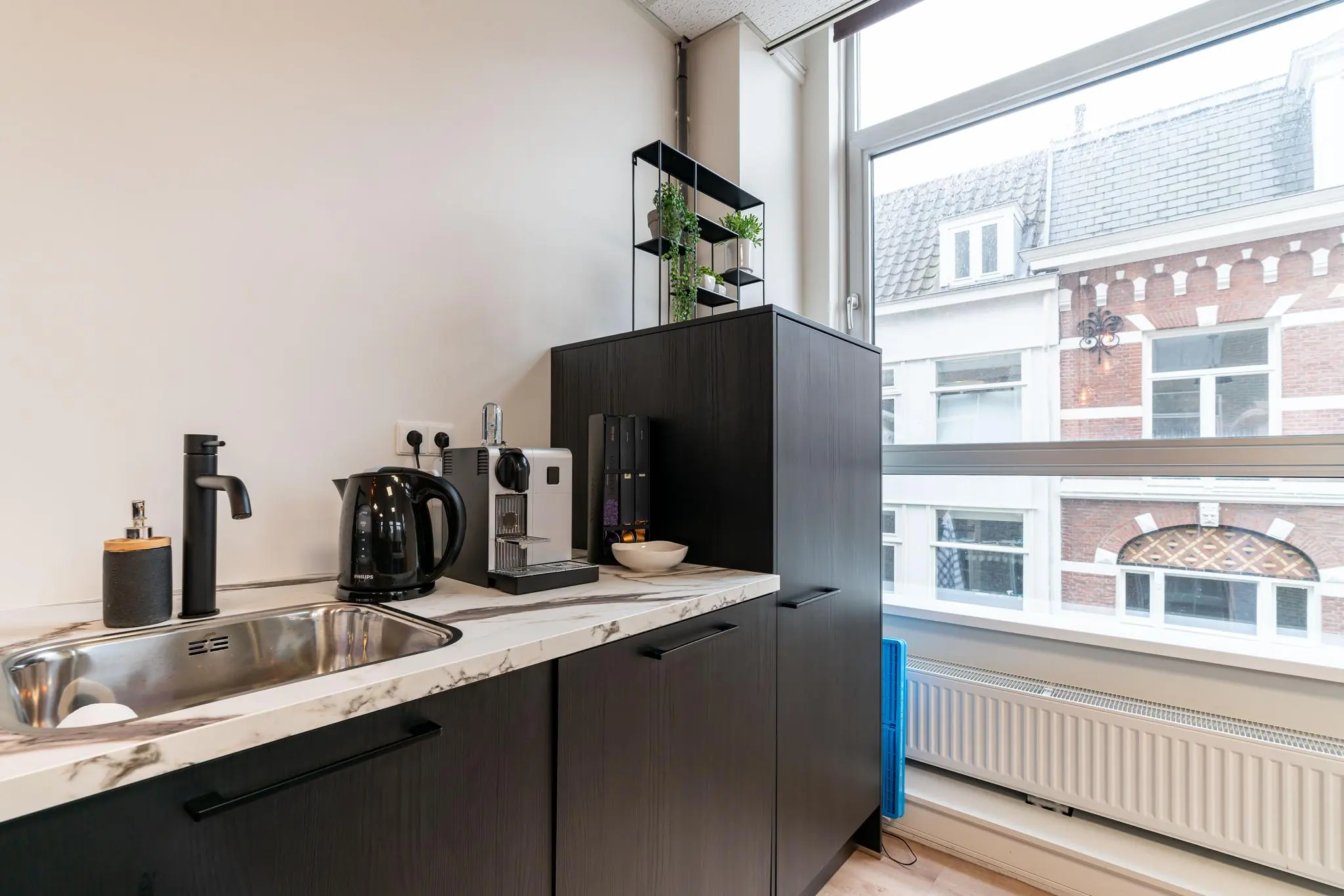 Moderne kitchenette met een koffiezetapparaat, waterkoker en spoelbak bij een groot raam met uitzicht op historische gevels aan het plein.