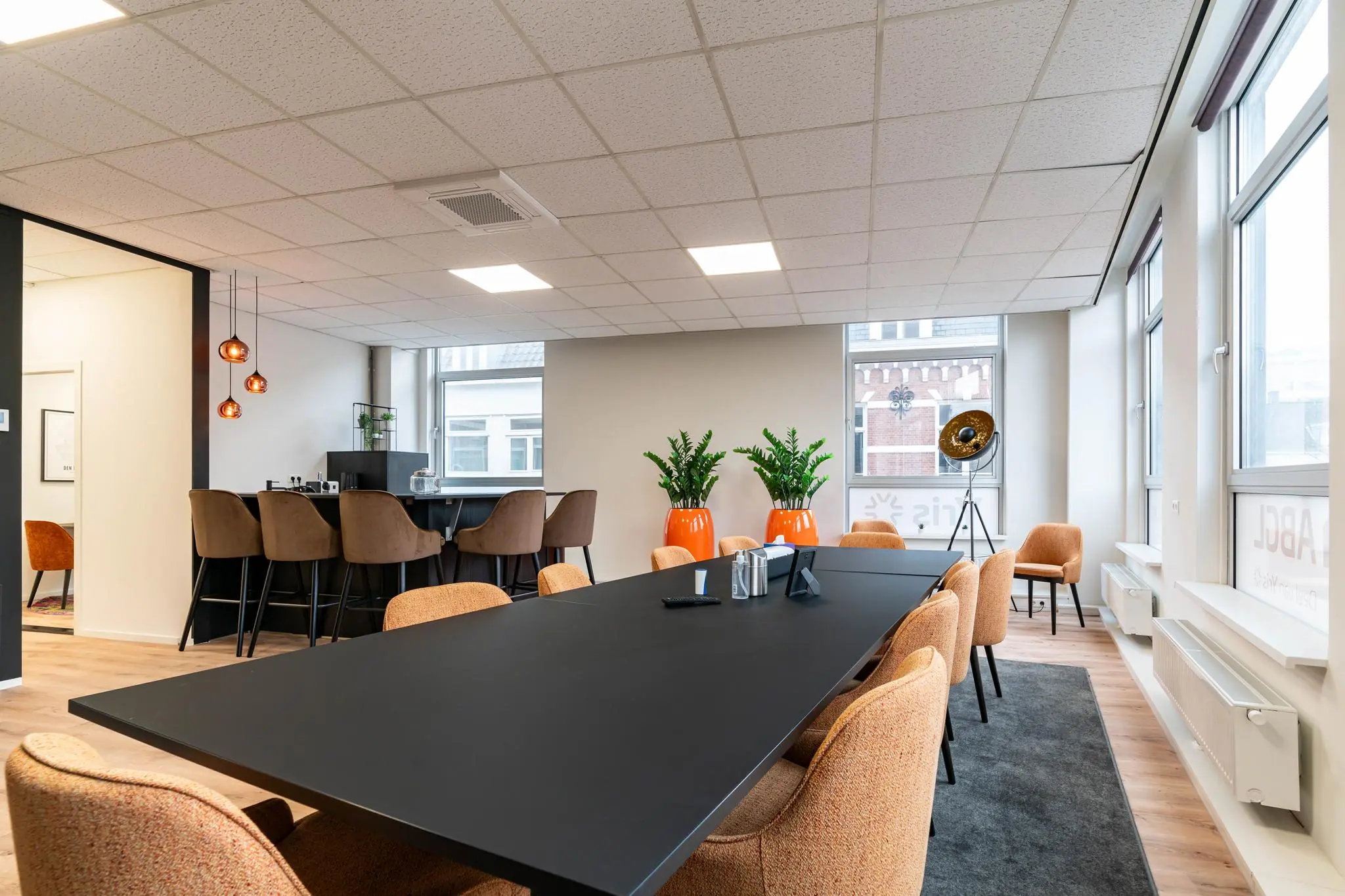 Moderne vergaderruimte met zwarte tafel, oranje stoelen, hoge zitplek bij de bar en grote ramen met uitzicht op het plein.