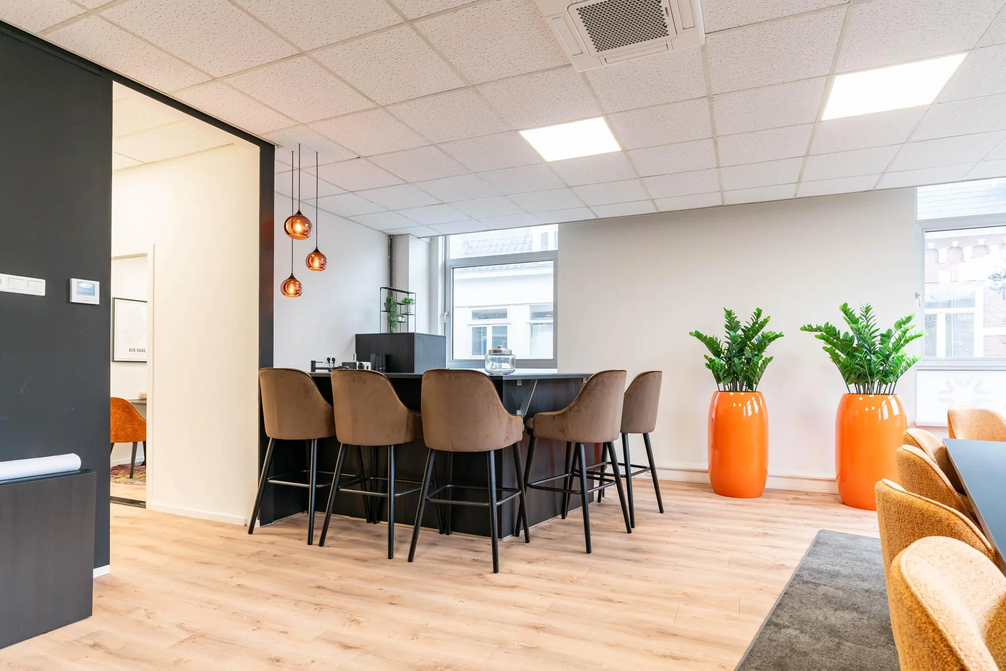 Moderne kantoorruimte met een hoge tafel, barkrukken, hanglampen en twee grote oranje plantenpotten bij het raam.