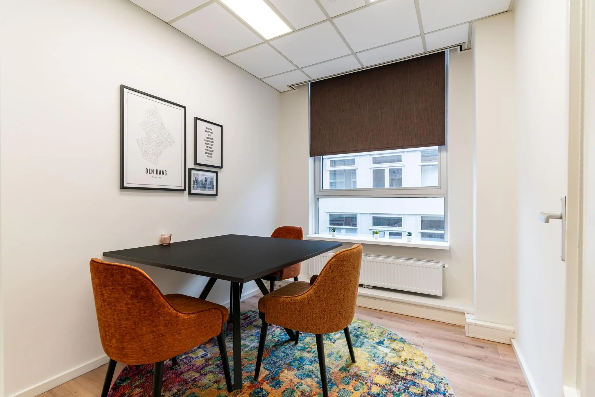 Kleine vergaderruimte met een zwarte tafel, drie oranje stoelen en een Den Haag-poster aan de muur.
