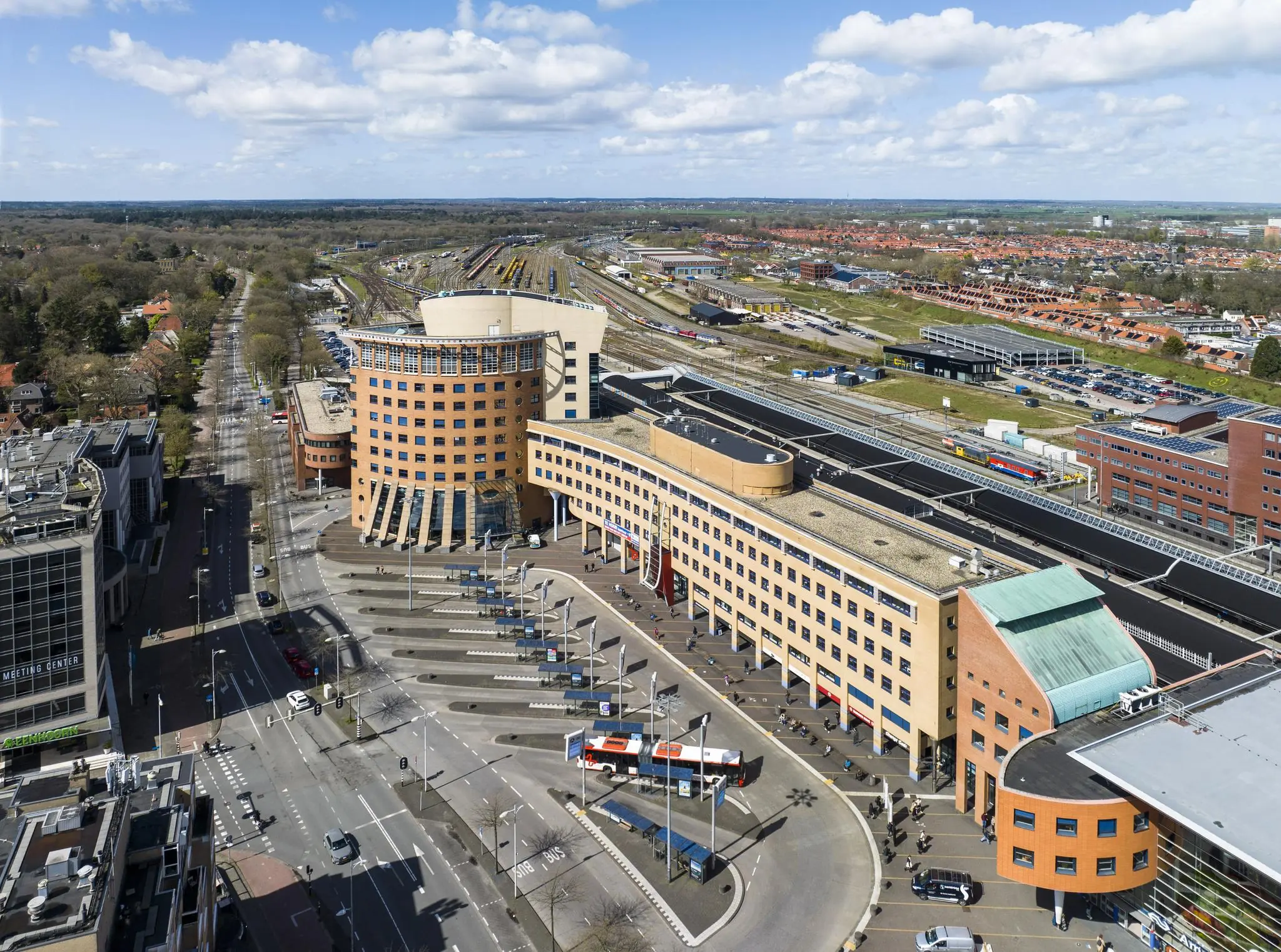 Luchtfoto van het Stationsplein in Enschede met het opvallende ronde stationsgebouw, busstation en treinsporen op de achtergrond.