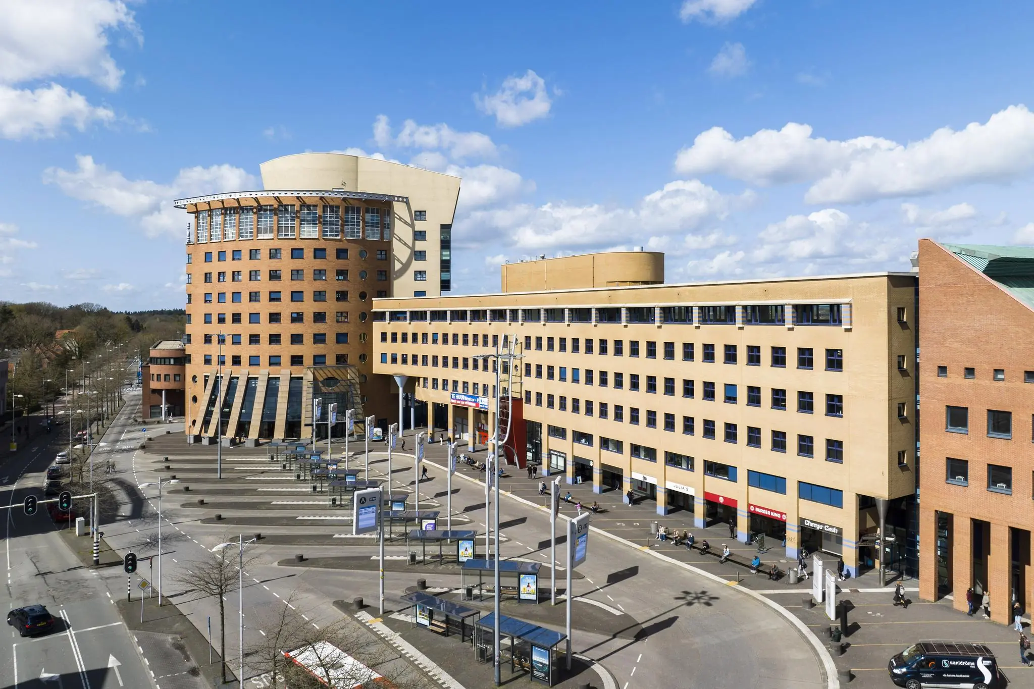 Modern kantoorgebouw aan het Stationsplein met bushaltes en voetgangers op een zonnige dag.